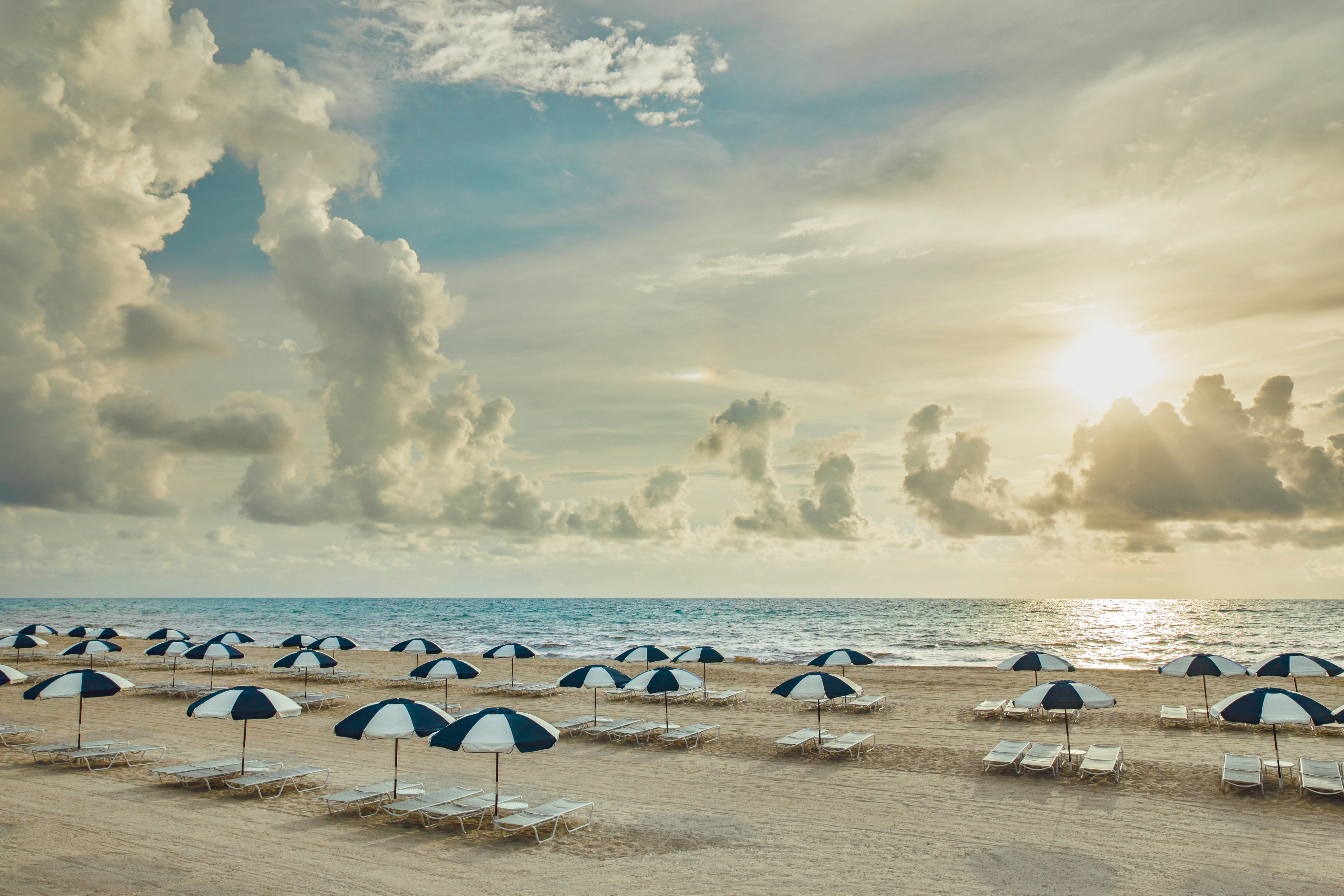 Beach scene with blue and white striped umbrellas and lounge chairs on sand under dramatic cloudy sky