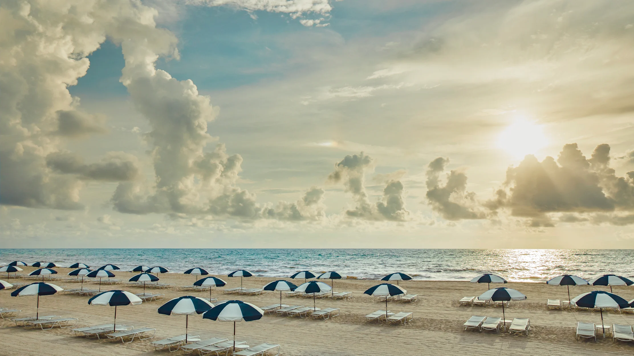 Beach scene with blue and white striped umbrellas and lounge chairs on sand under dramatic cloudy sky