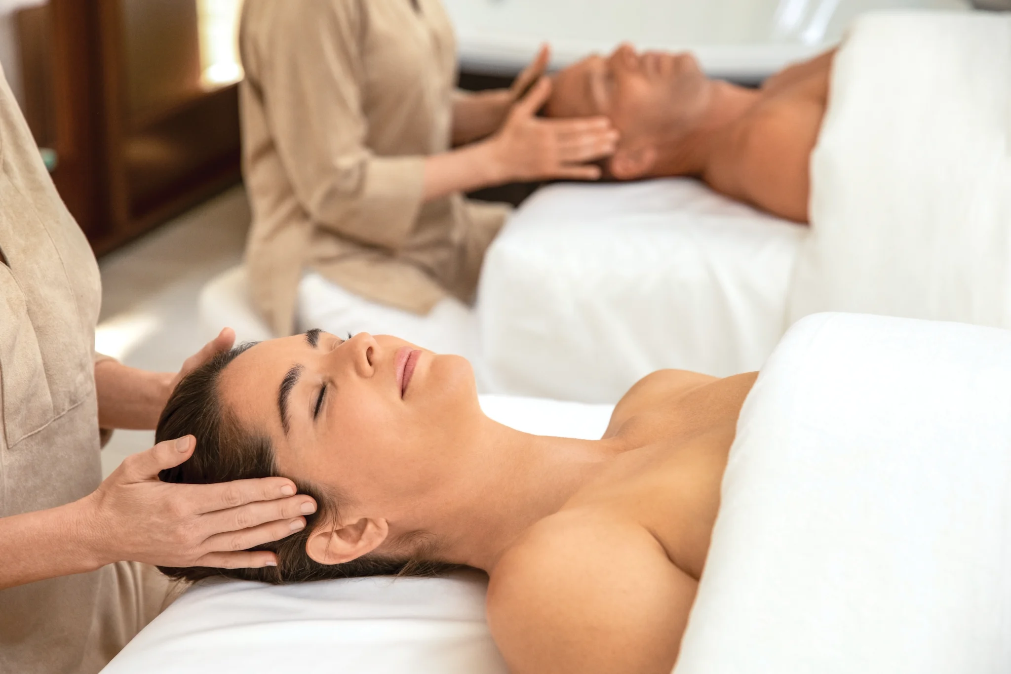 Two people receiving relaxing head massages at a spa, lying on white massage tables.