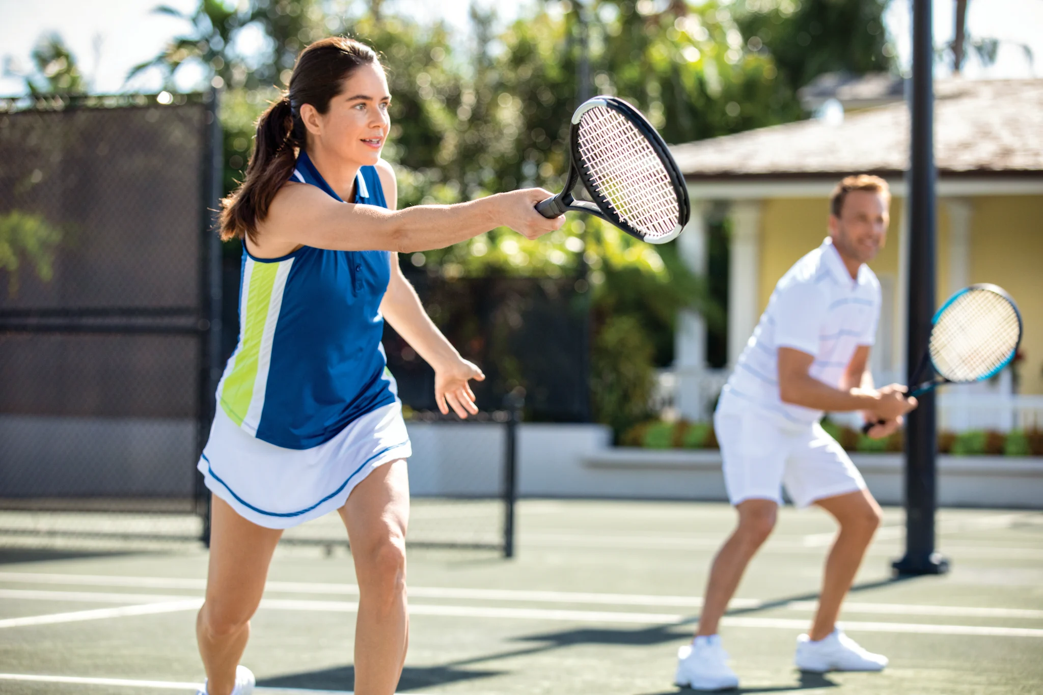Woman in blue and white tennis outfit prepares to hit ball on sunny outdoor court with male partner.