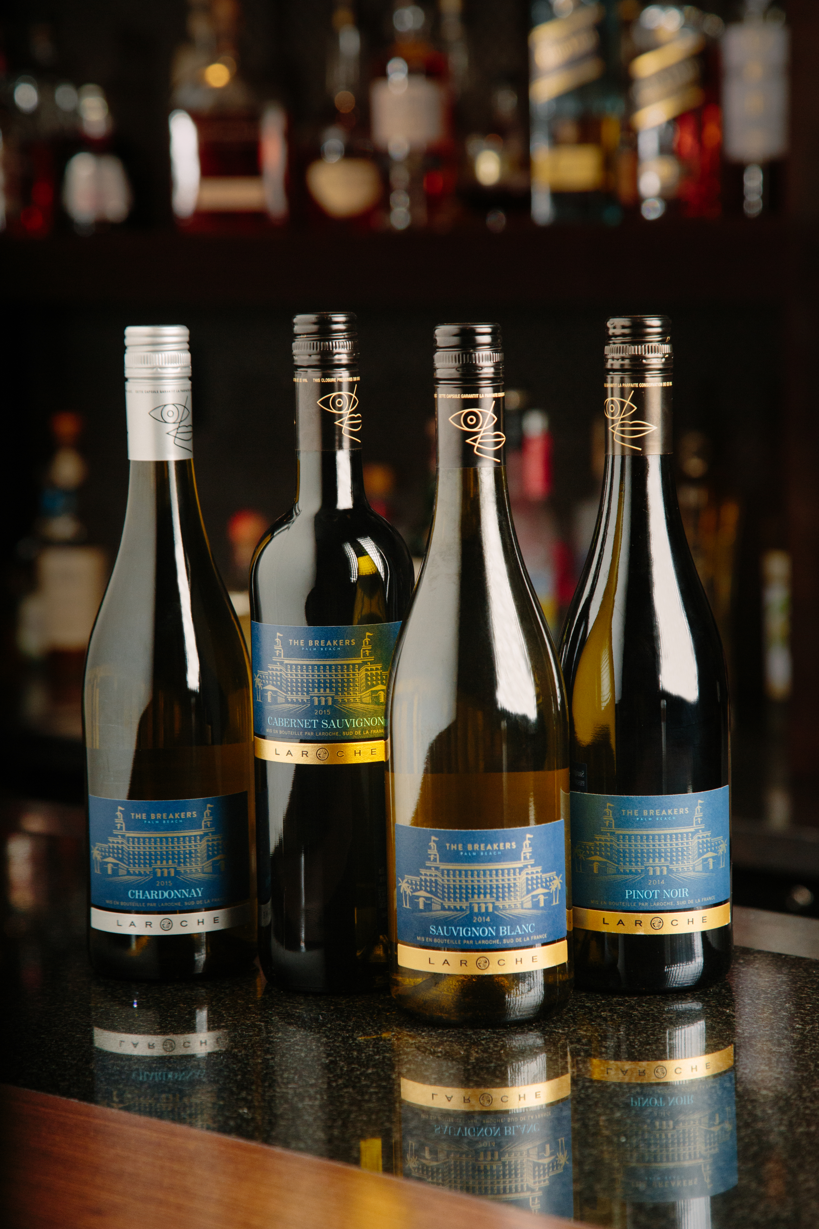 Four wine bottles with blue and gold labels displayed on a reflective bar surface with blurred bottles in the background.
