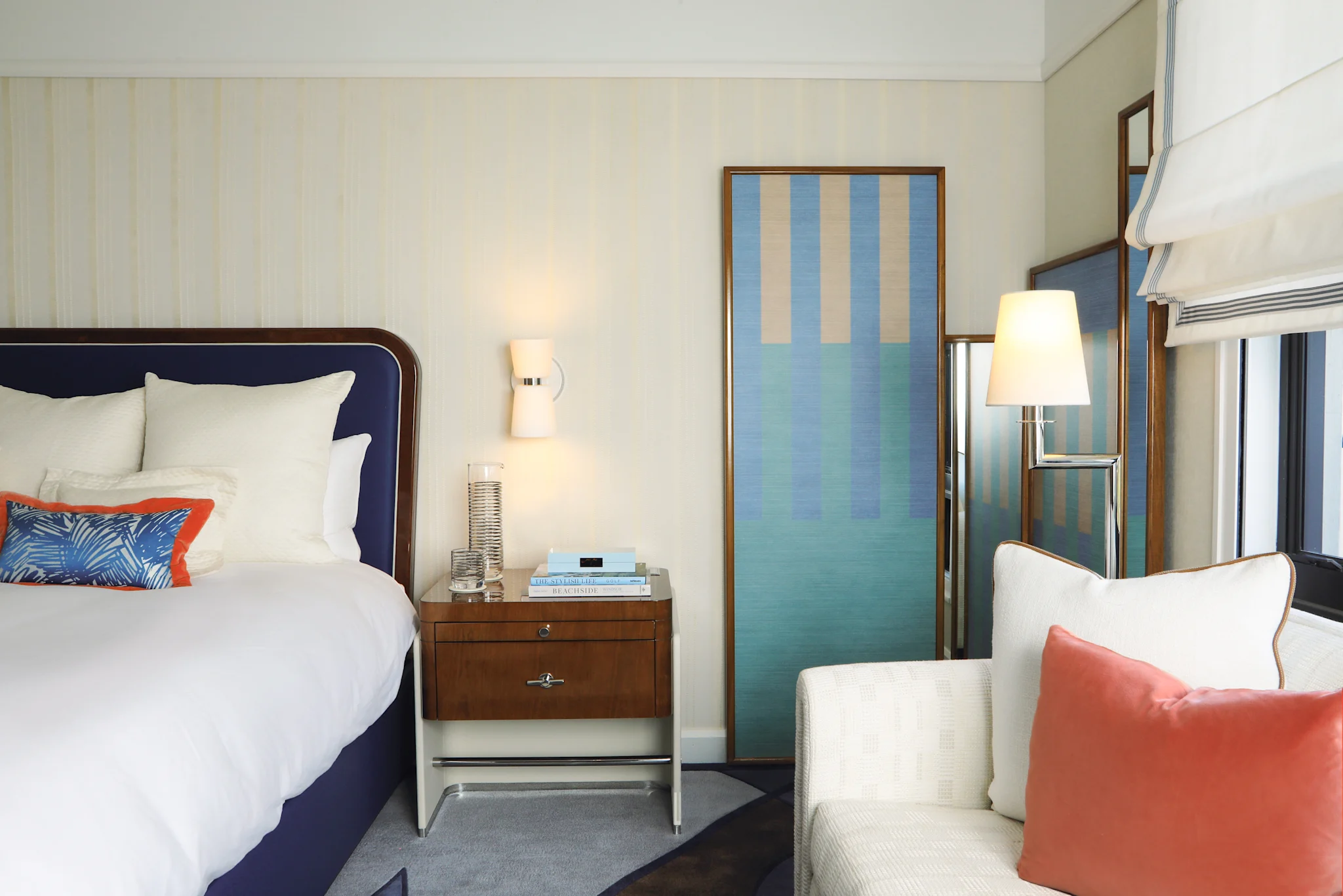 Modern hotel bedroom with navy upholstered bed, white chair, coral pillows, and blue striped artwork.