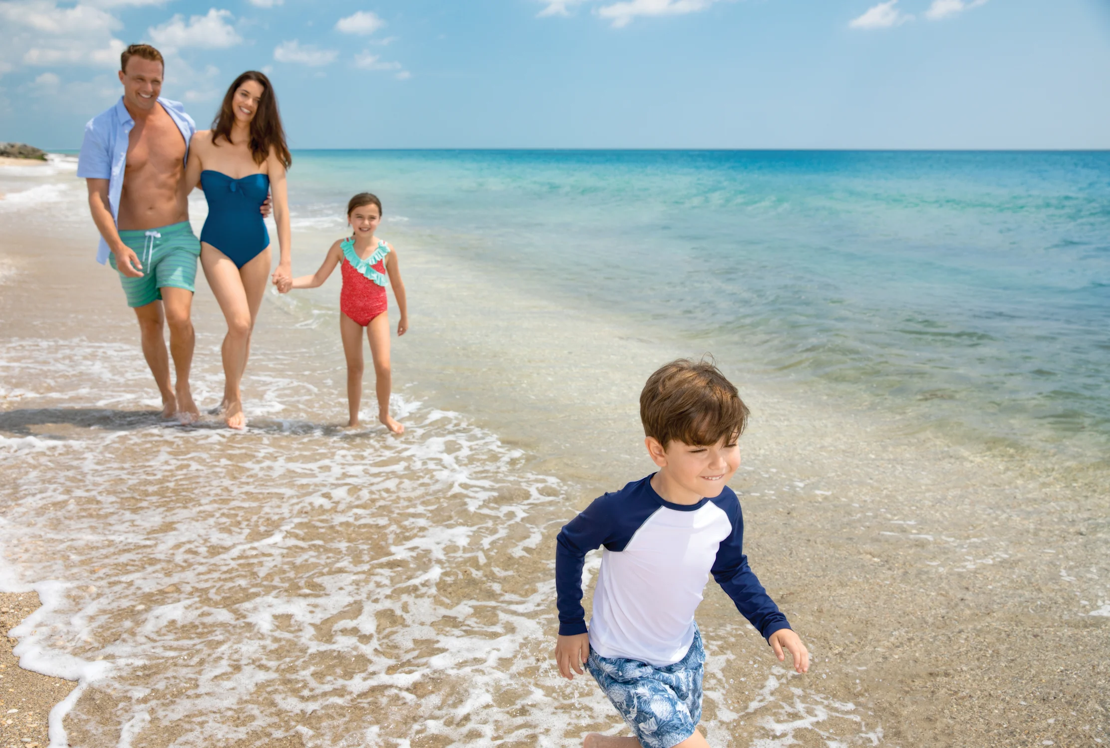 Happy family of four walking on a sunny beach with turquoise water and blue sky.