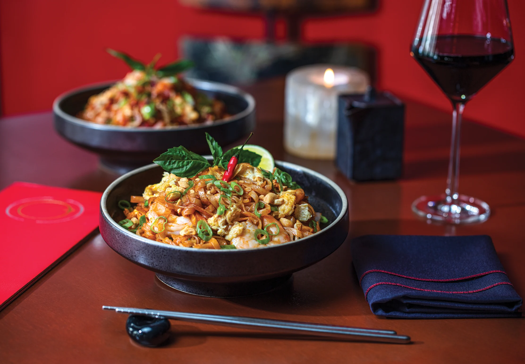 Two black bowls of Asian noodle dishes with vegetables and herbs, served with chopsticks and red wine.