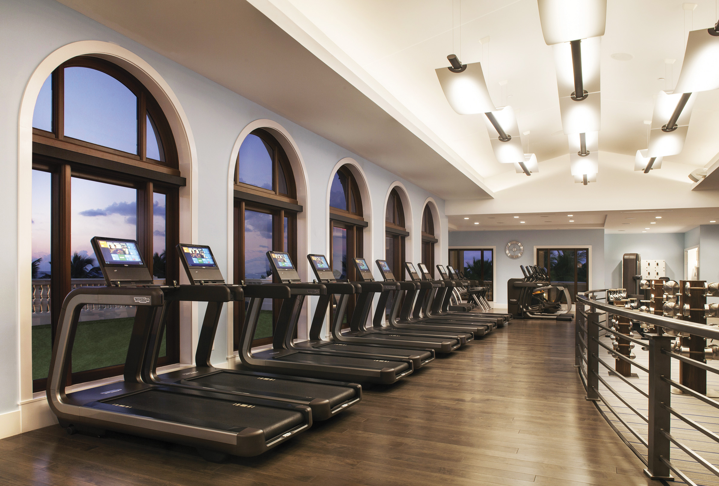 Modern fitness center with row of treadmills along arched windows, wood floors, and weight racks.