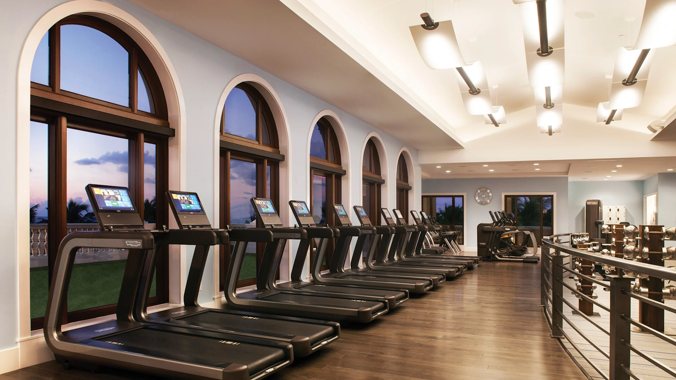 Modern fitness center with row of treadmills along arched windows, wood floors, and weight racks.