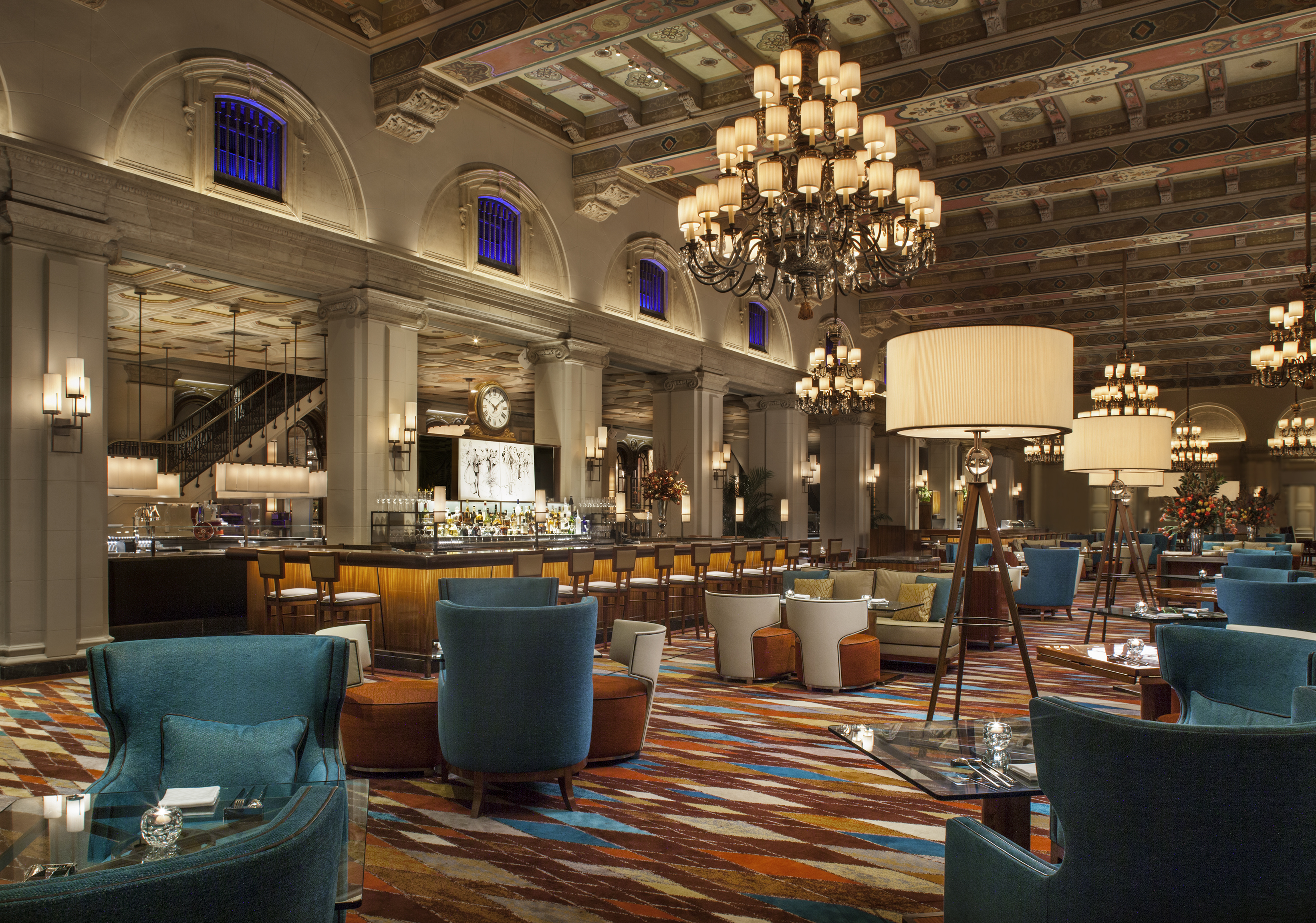 Elegant hotel lobby with ornate ceiling, chandeliers, bar area, and teal and orange seating arrangements.