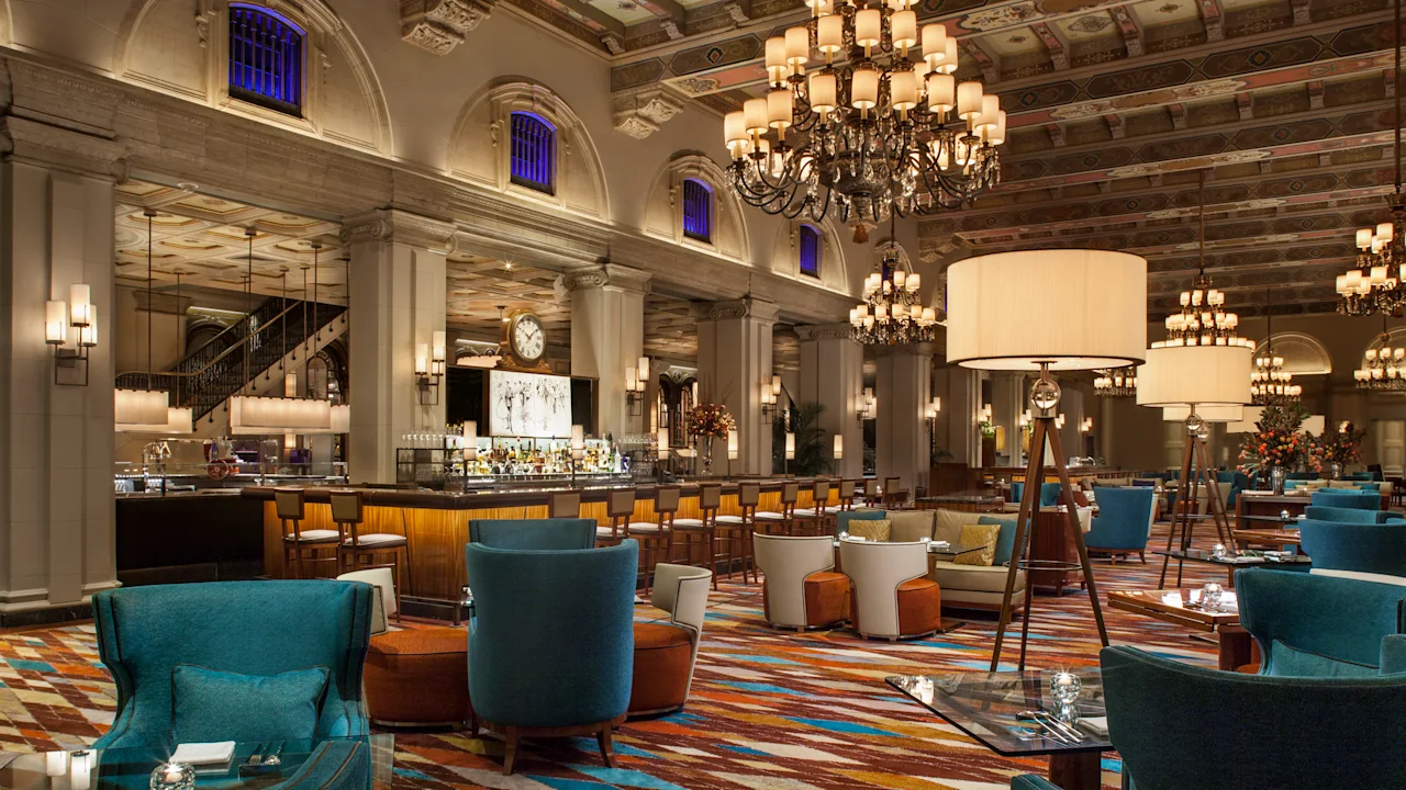 Elegant hotel lobby with ornate ceiling, chandeliers, bar area, and teal and orange seating arrangements.