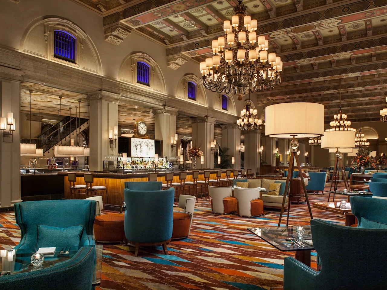 Elegant hotel restaurant with ornate ceiling, chandeliers, bar and teal and orange seating areas.
