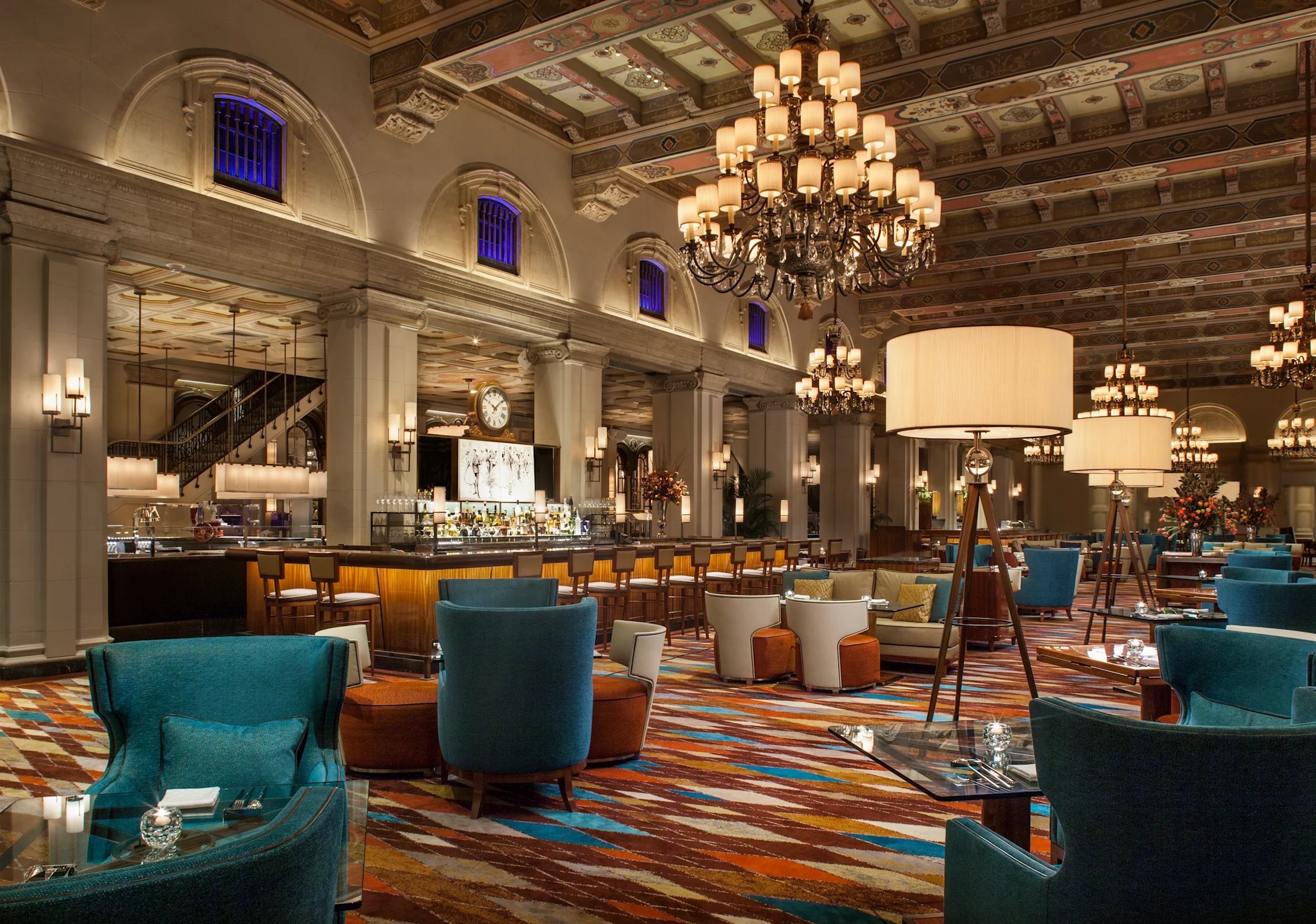 Elegant hotel lobby with ornate ceiling, chandeliers, bar area, and teal and orange seating arrangements.