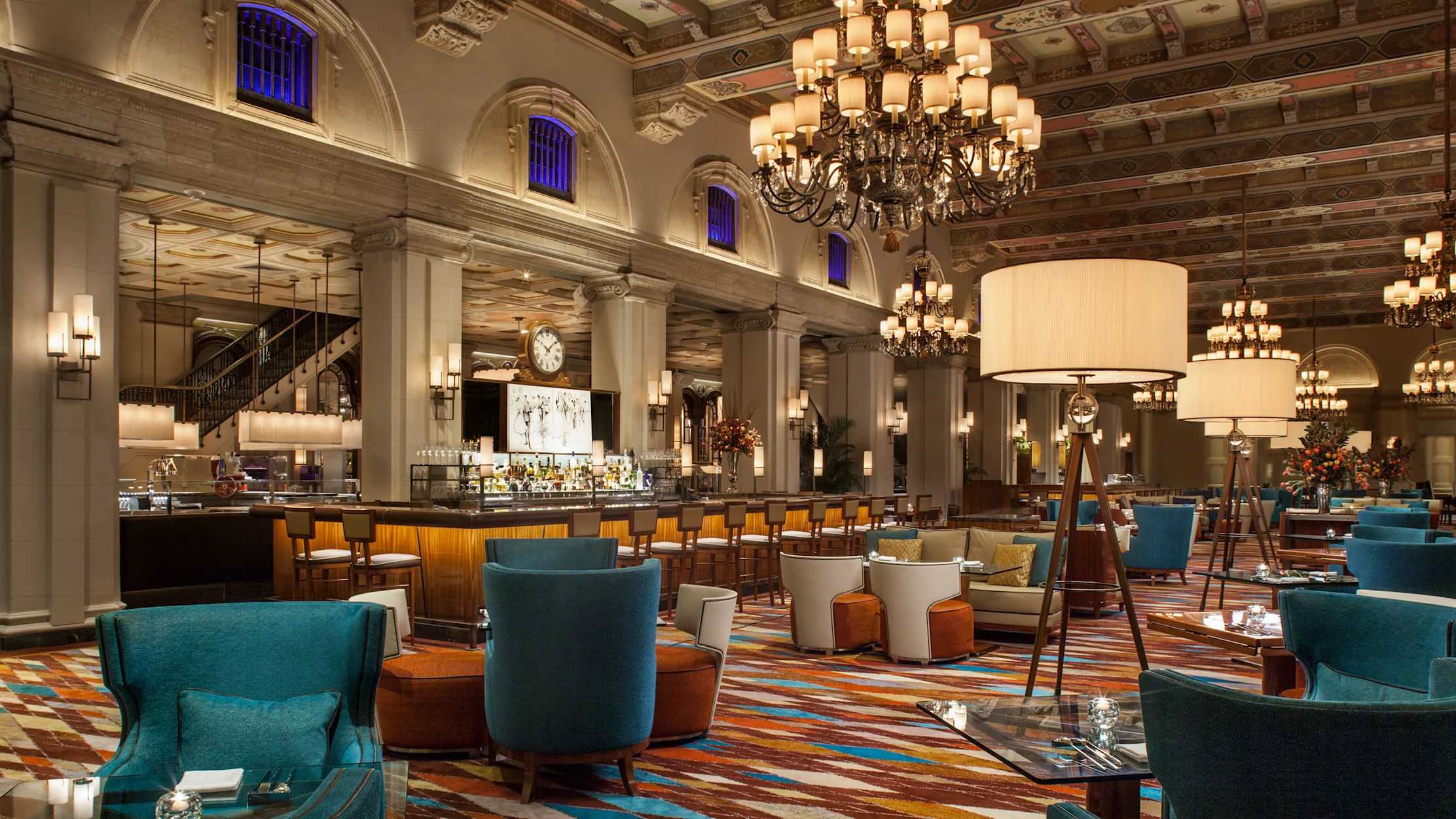 Elegant hotel restaurant with ornate ceiling, chandeliers, bar and teal and orange seating areas.