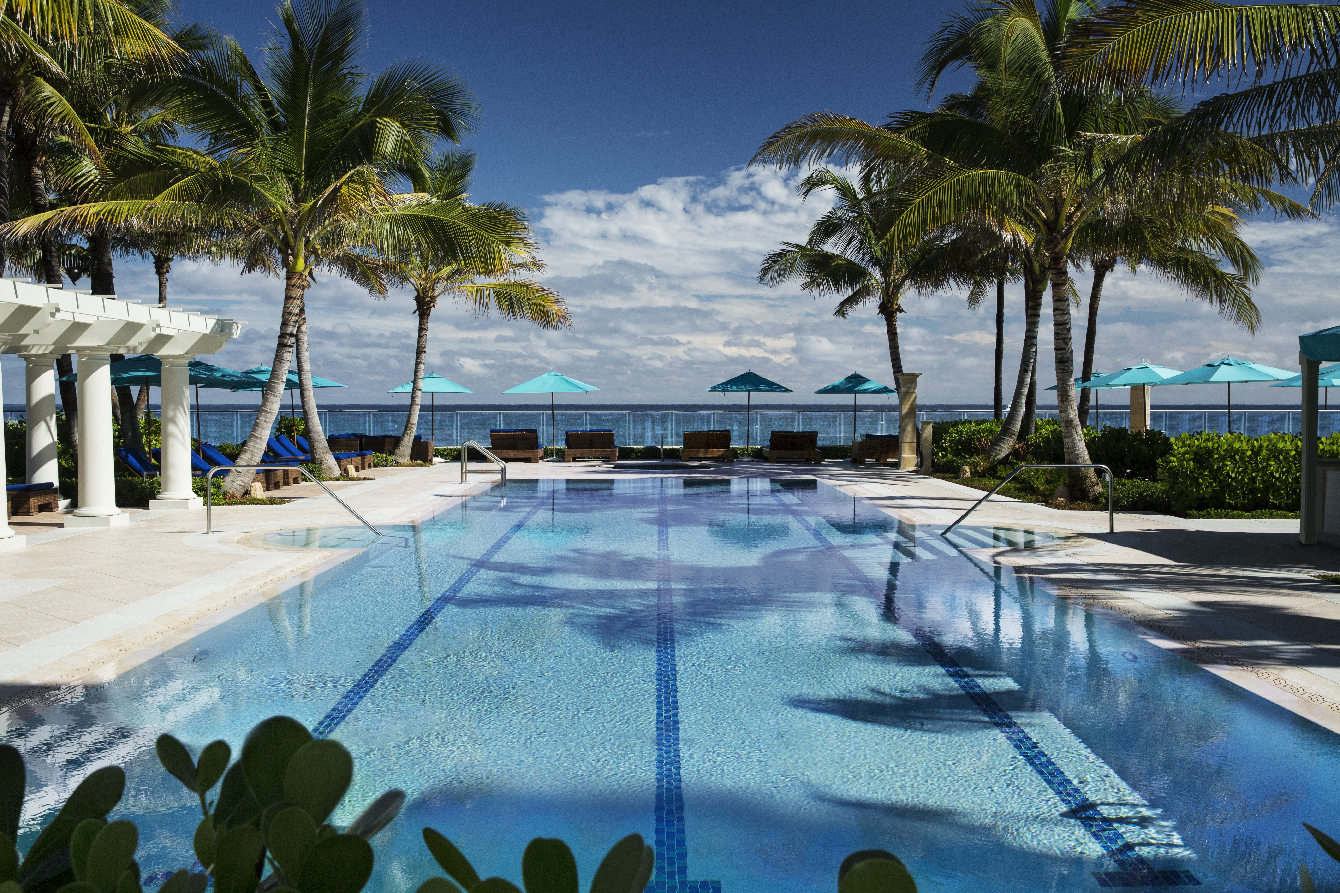 Tropical resort pool with palm trees, turquoise umbrellas, and ocean view under blue sky.