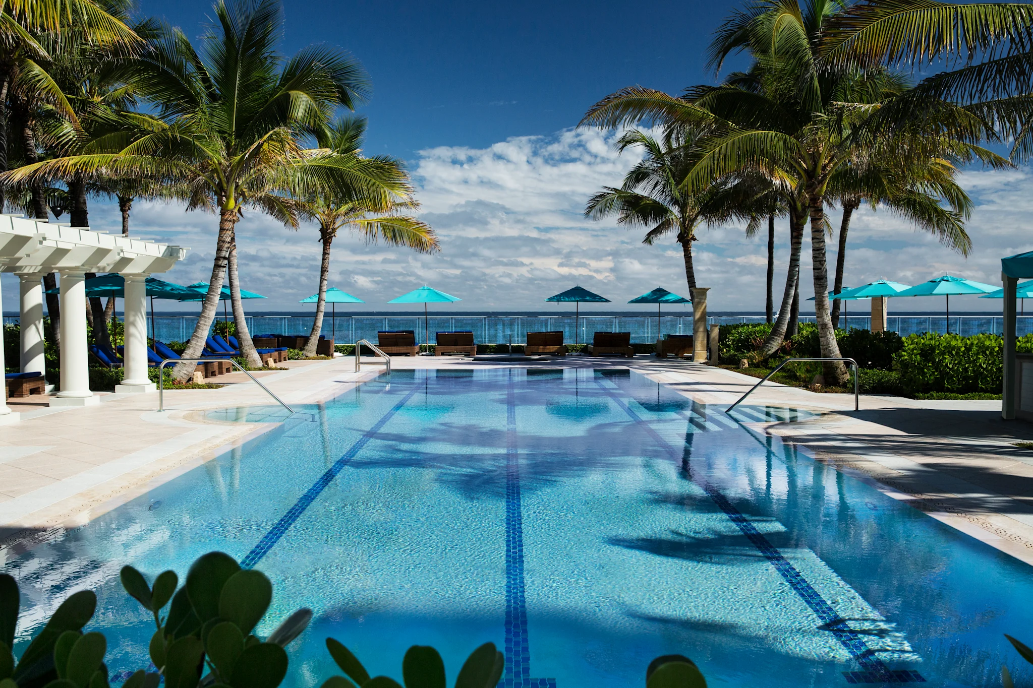 Tropical resort pool with palm trees, turquoise umbrellas, and ocean view under blue sky.