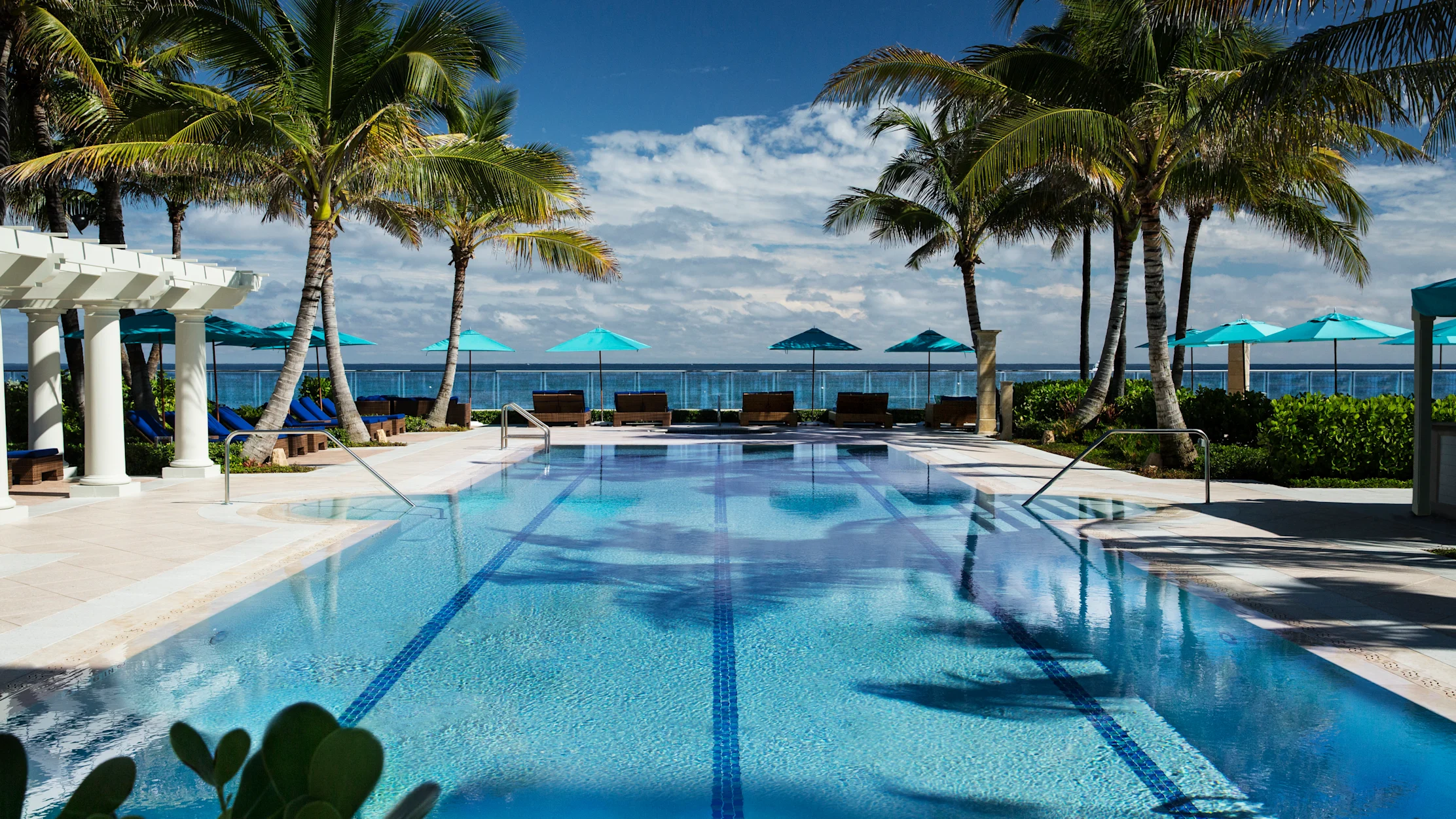 Tropical resort pool with palm trees, turquoise umbrellas, and ocean view under blue sky.