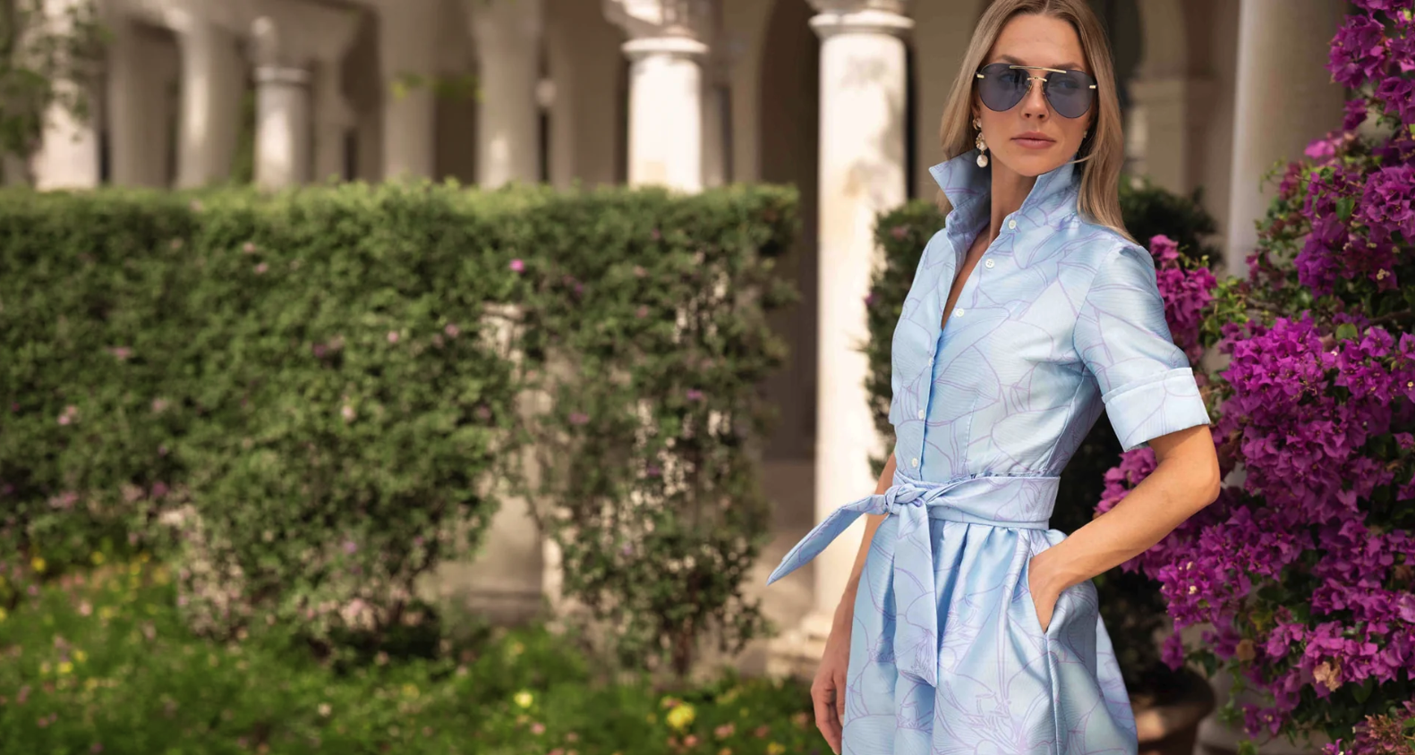 Woman in light blue dress and sunglasses standing near purple flowers and green hedges outdoors.