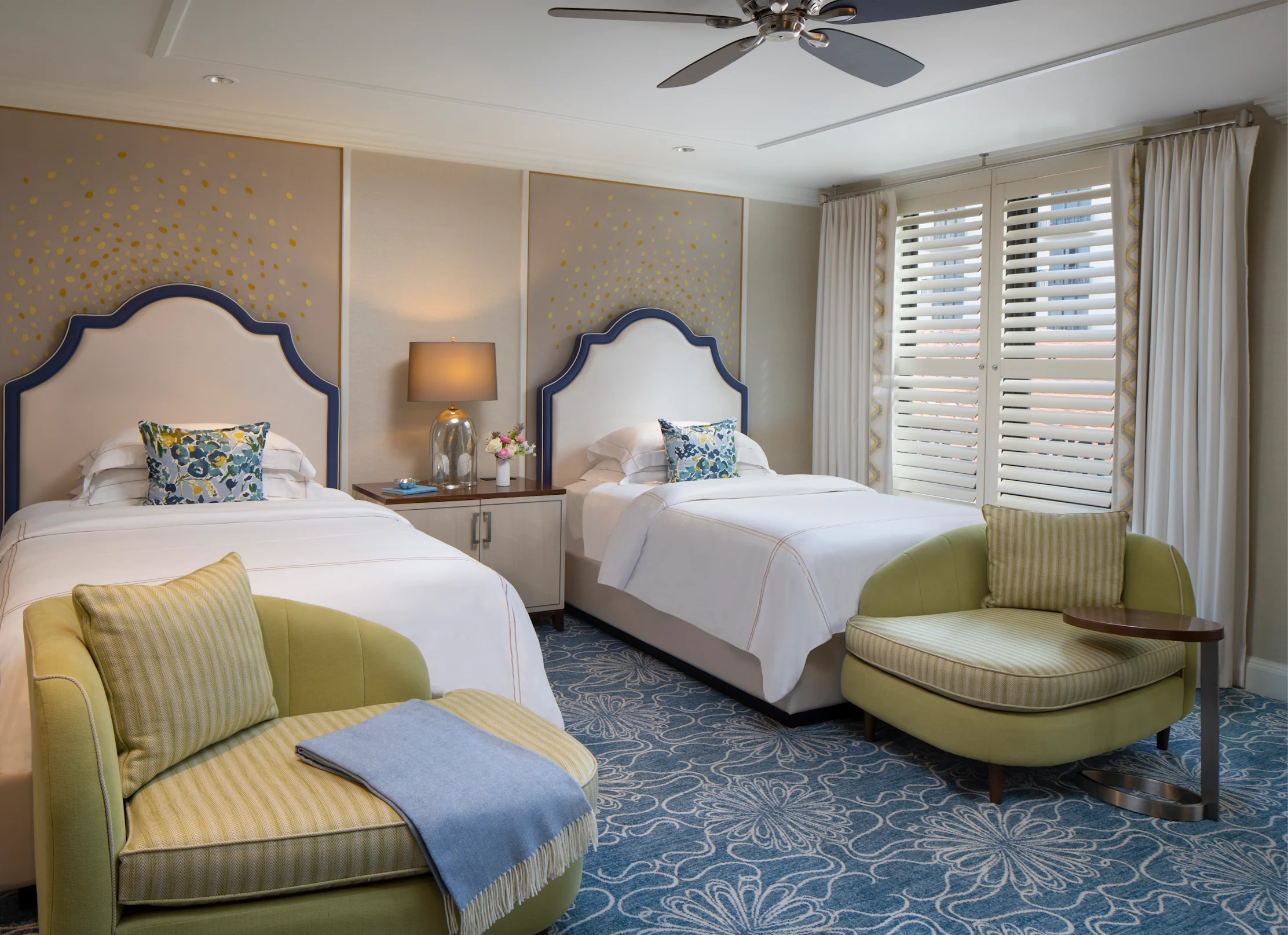 Elegant hotel room with two twin beds with navy headboards, green armchairs, and gold dot wallpaper.