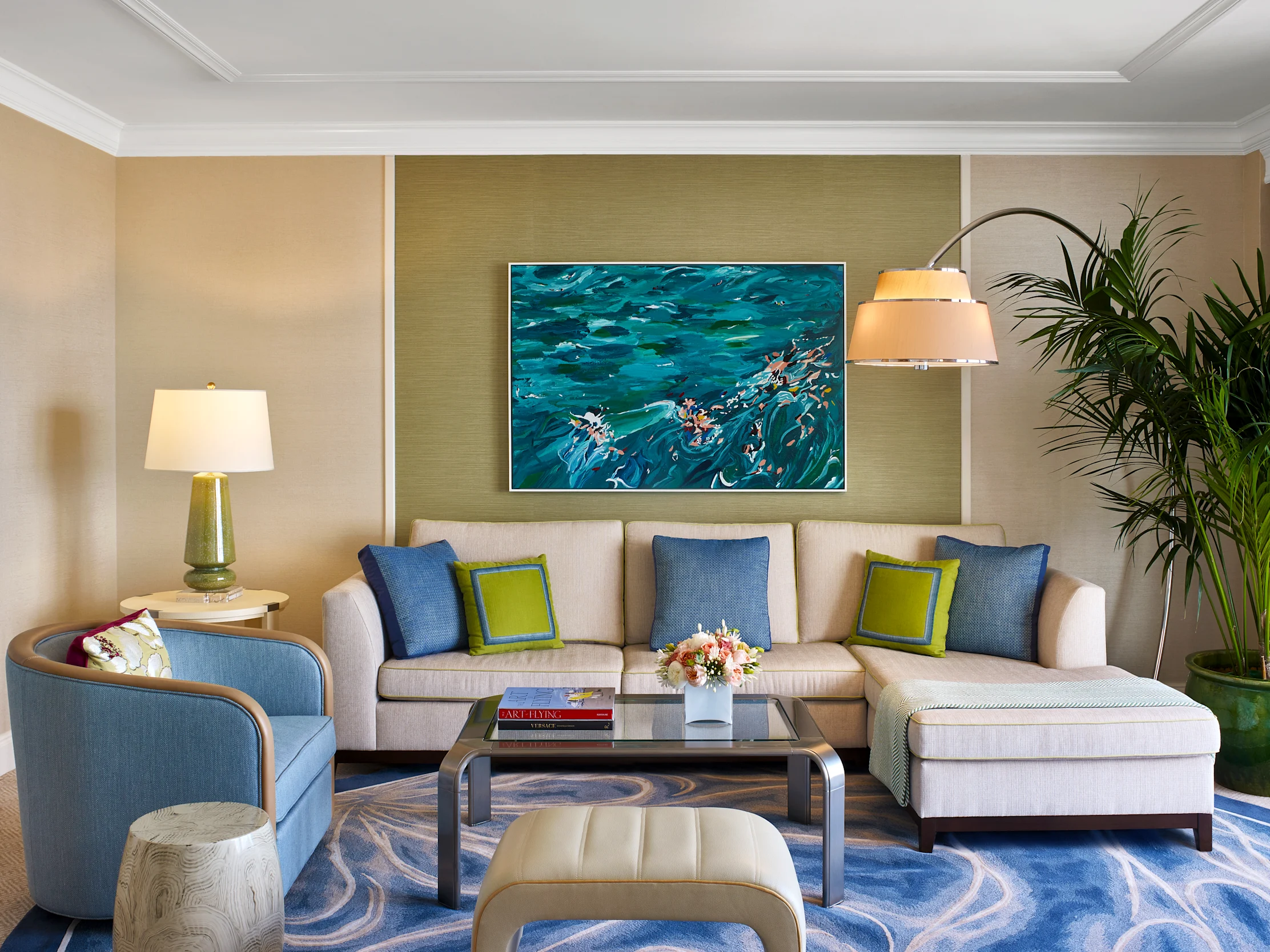 Modern living room with beige sectional sofa, blue ocean painting, green accent wall, and palm plants.