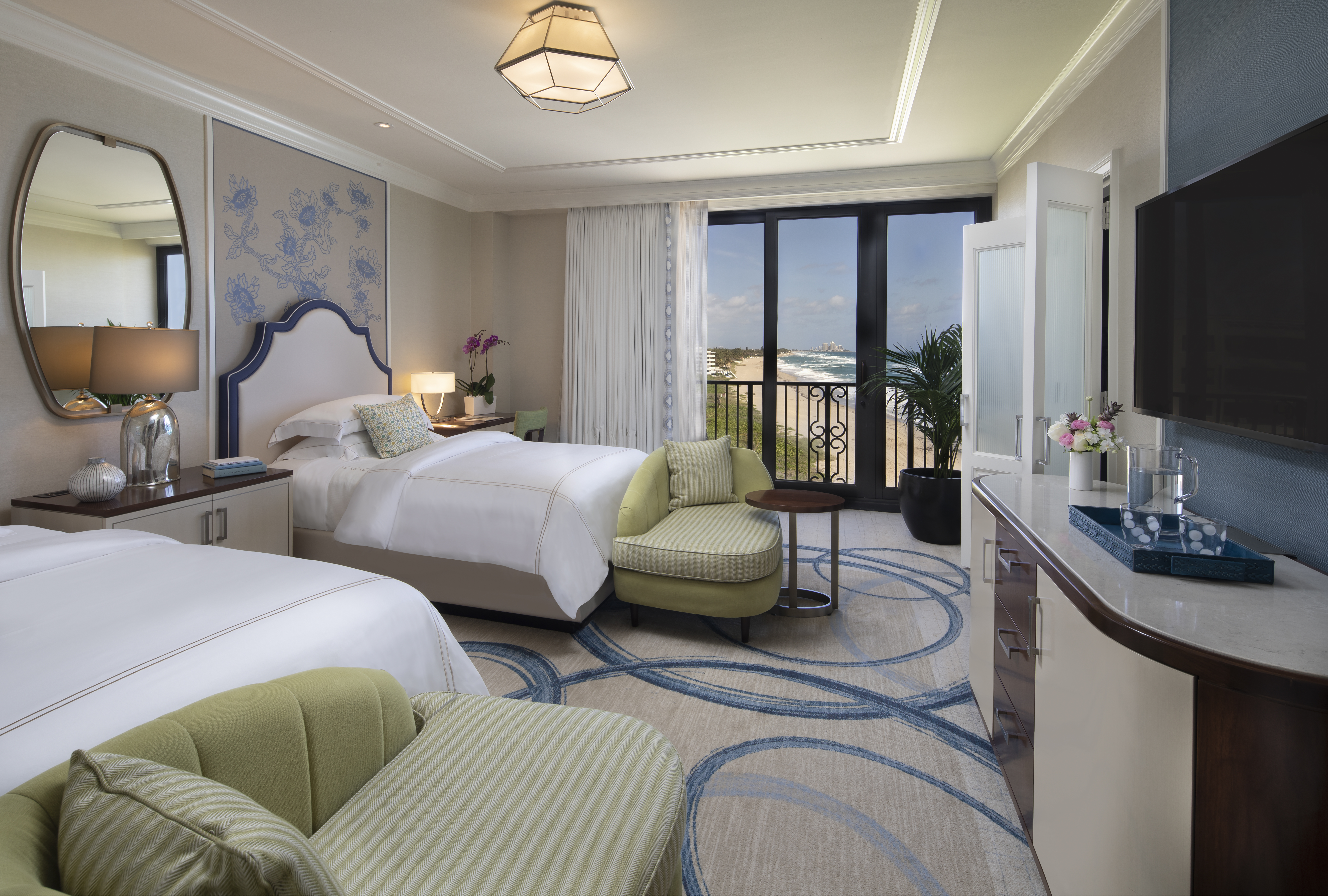 Elegant hotel room with two beds, green accent chairs, ocean view balcony, and modern decor.