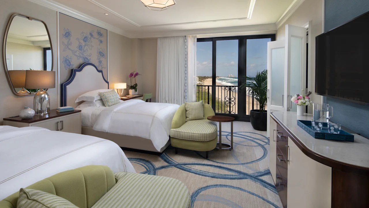 Elegant hotel room with two beds, green accent chairs, ocean view balcony, and modern decor.