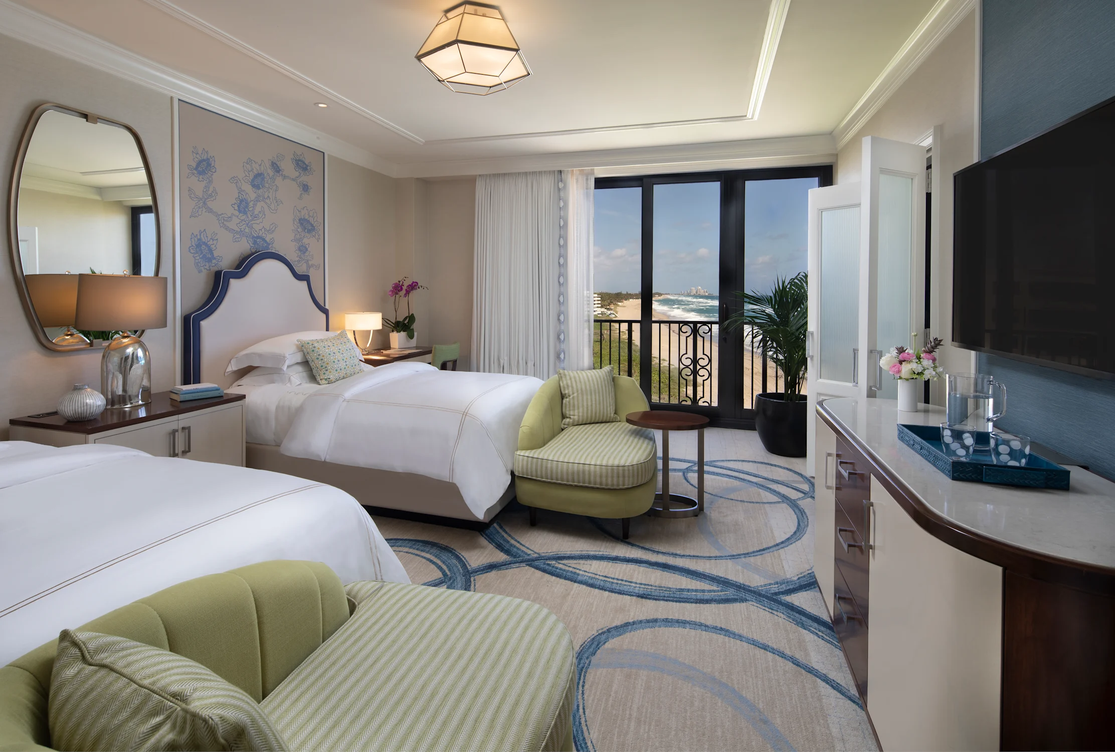 Elegant hotel room with two beds, green accent chairs, ocean view balcony, and modern decor.