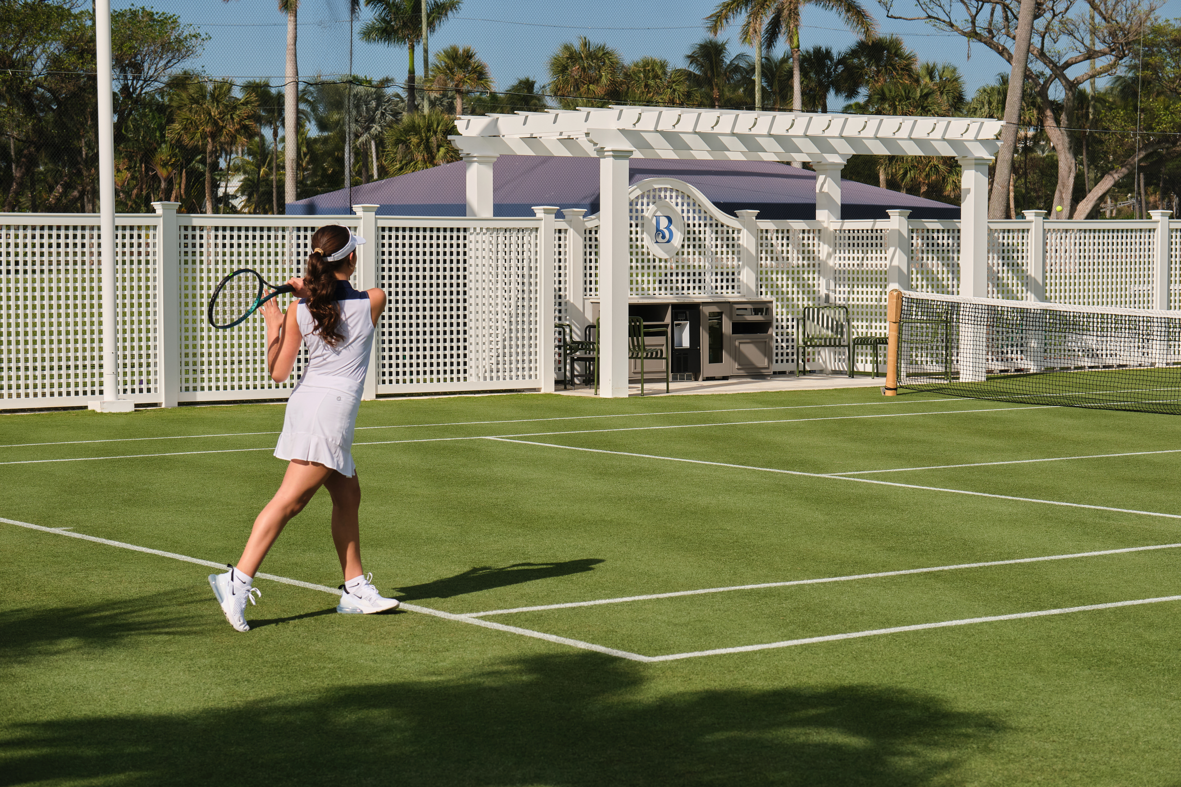 Woman in white tennis outfit playing on green court with white pergola and palm trees in background.