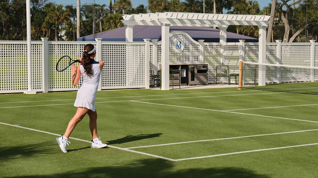 Woman in white tennis outfit playing on green court with white pergola and palm trees in background.