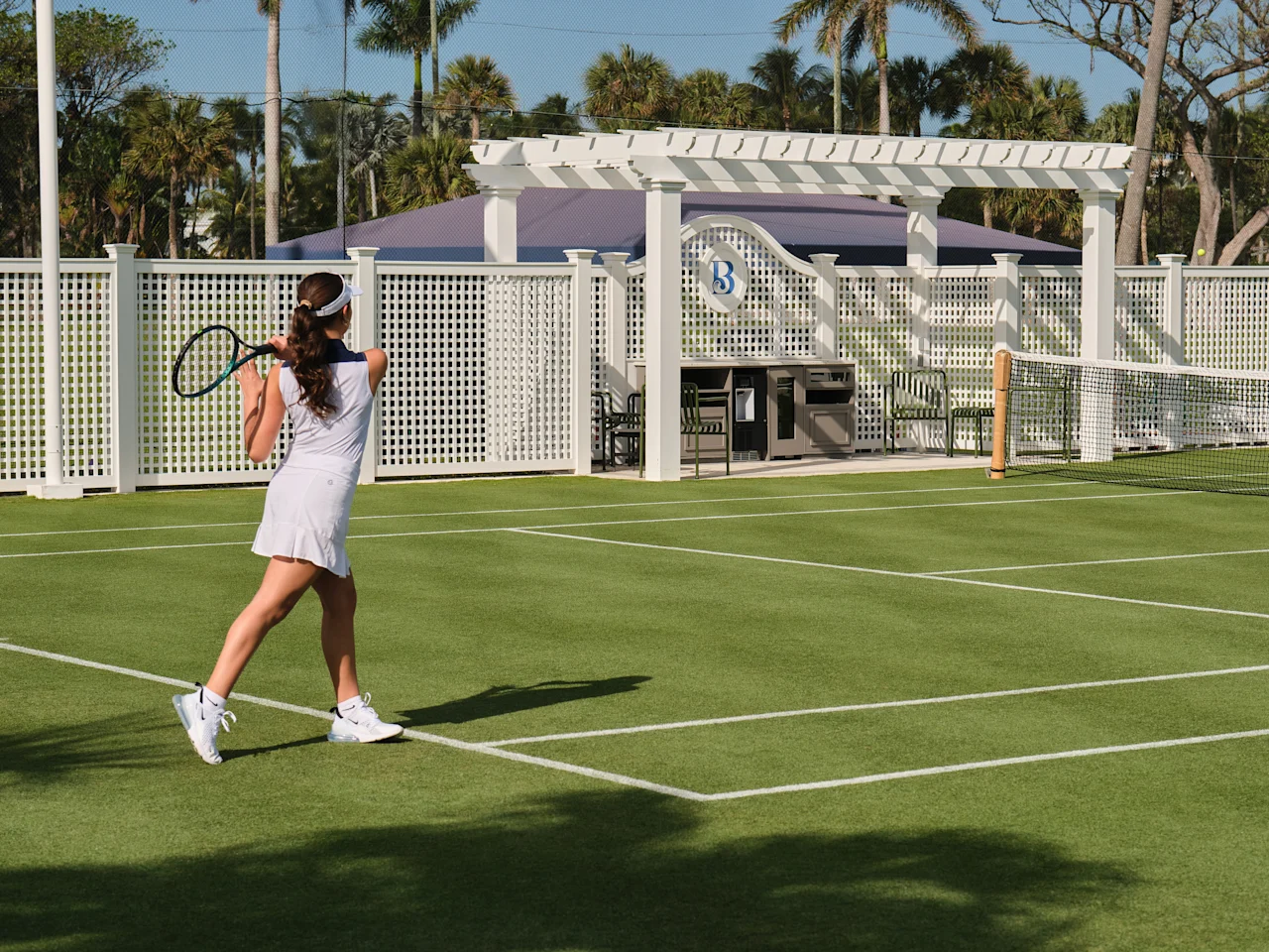Woman in white tennis outfit playing on green court with white pergola and palm trees in background.