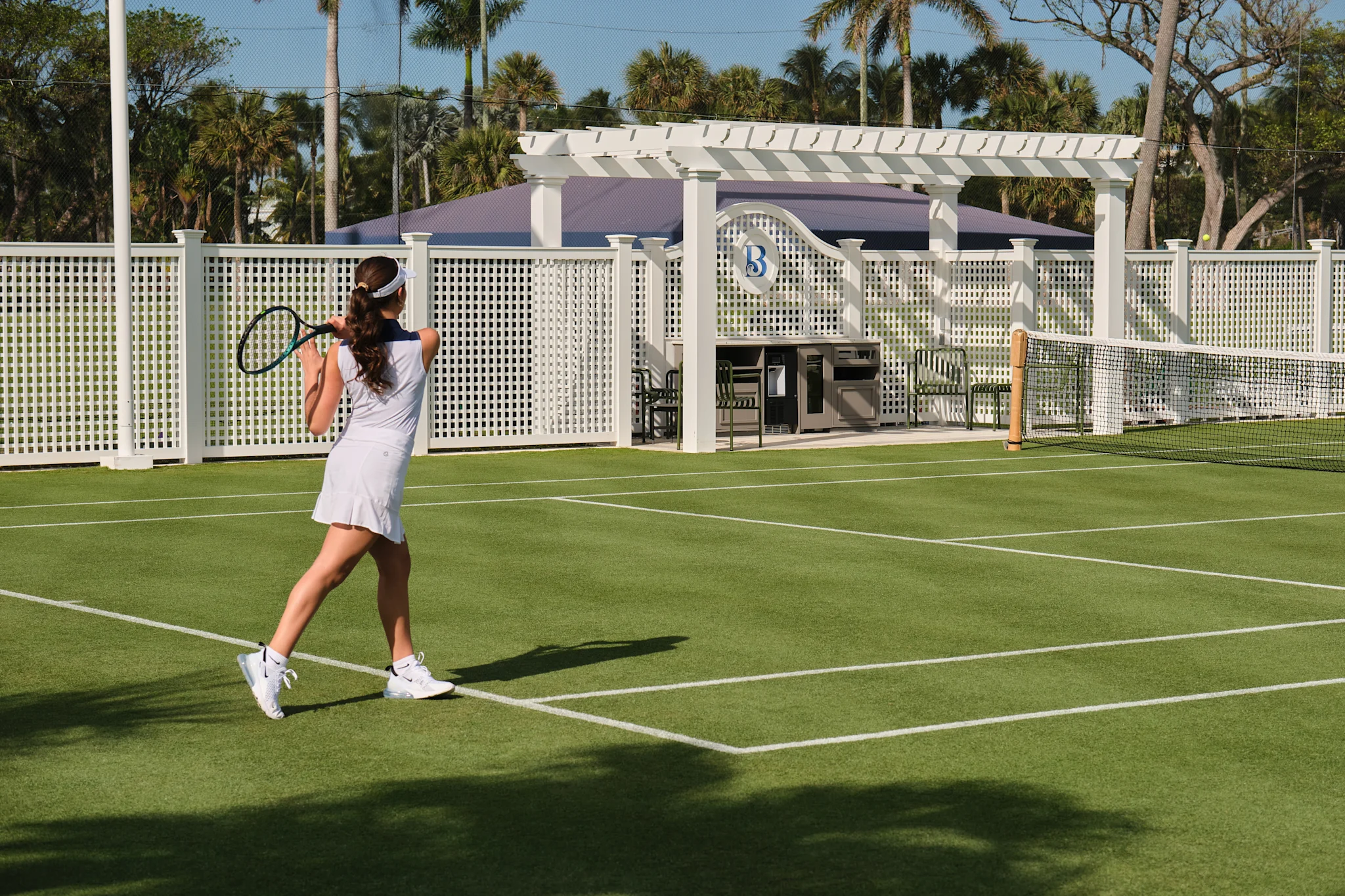 Woman in white tennis outfit playing on green court with white pergola and palm trees in background.