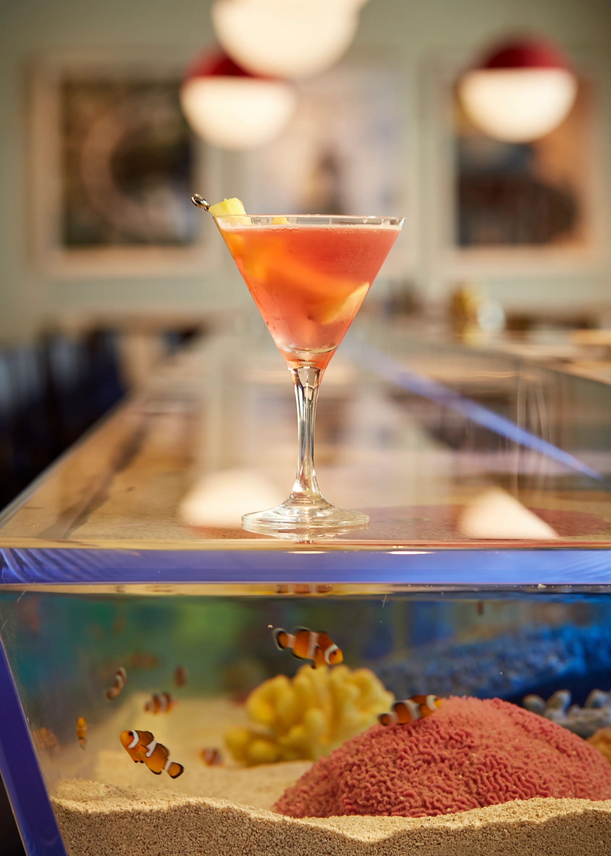 Orange cocktail in martini glass with lemon twist, displayed on aquarium with clownfish below.