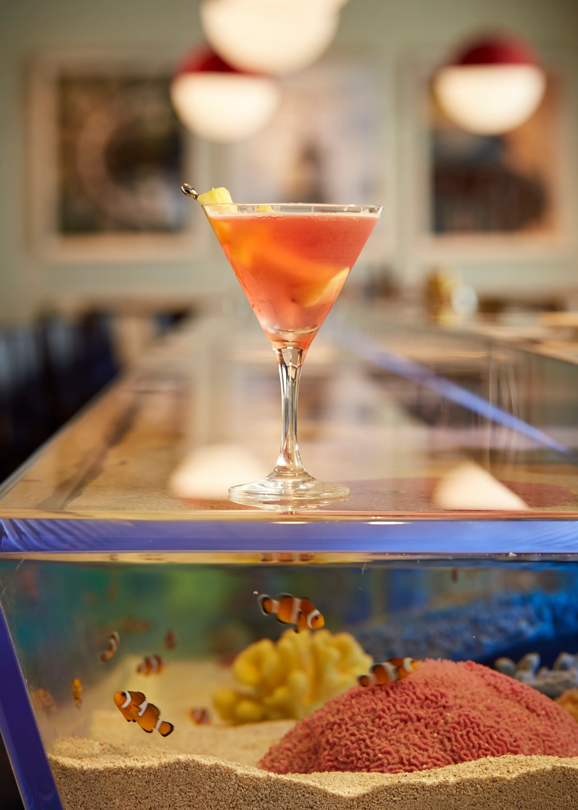 Orange cocktail in martini glass with lemon twist, displayed on aquarium with clownfish below.