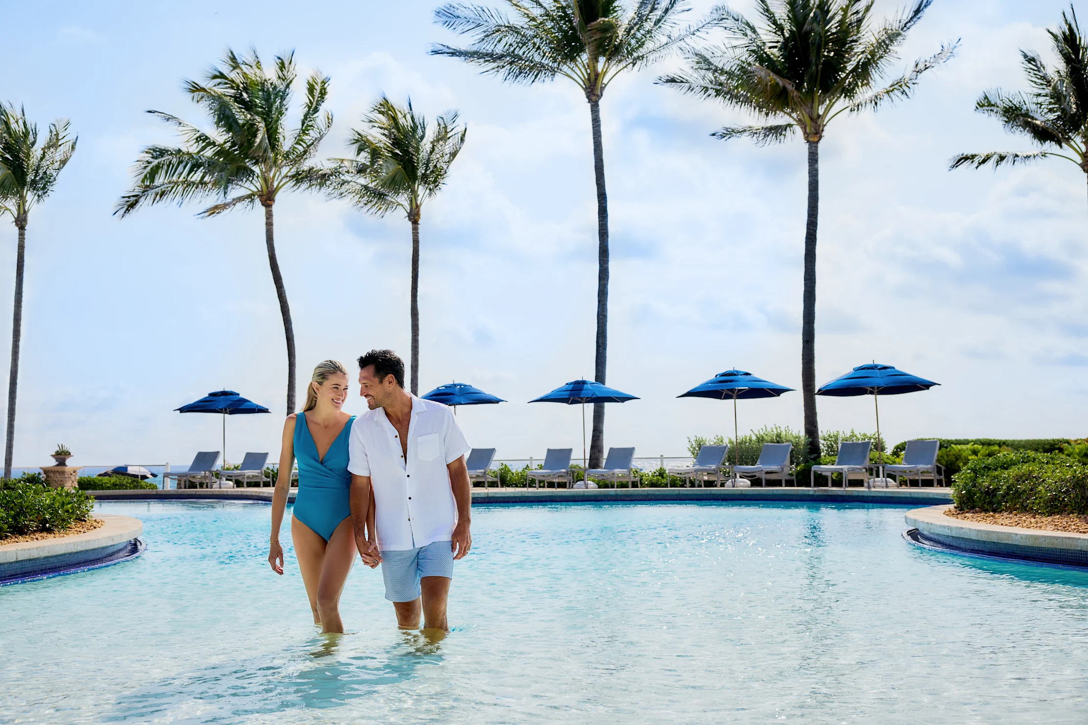 Couple walking in tropical resort pool with palm trees and blue umbrellas in background.