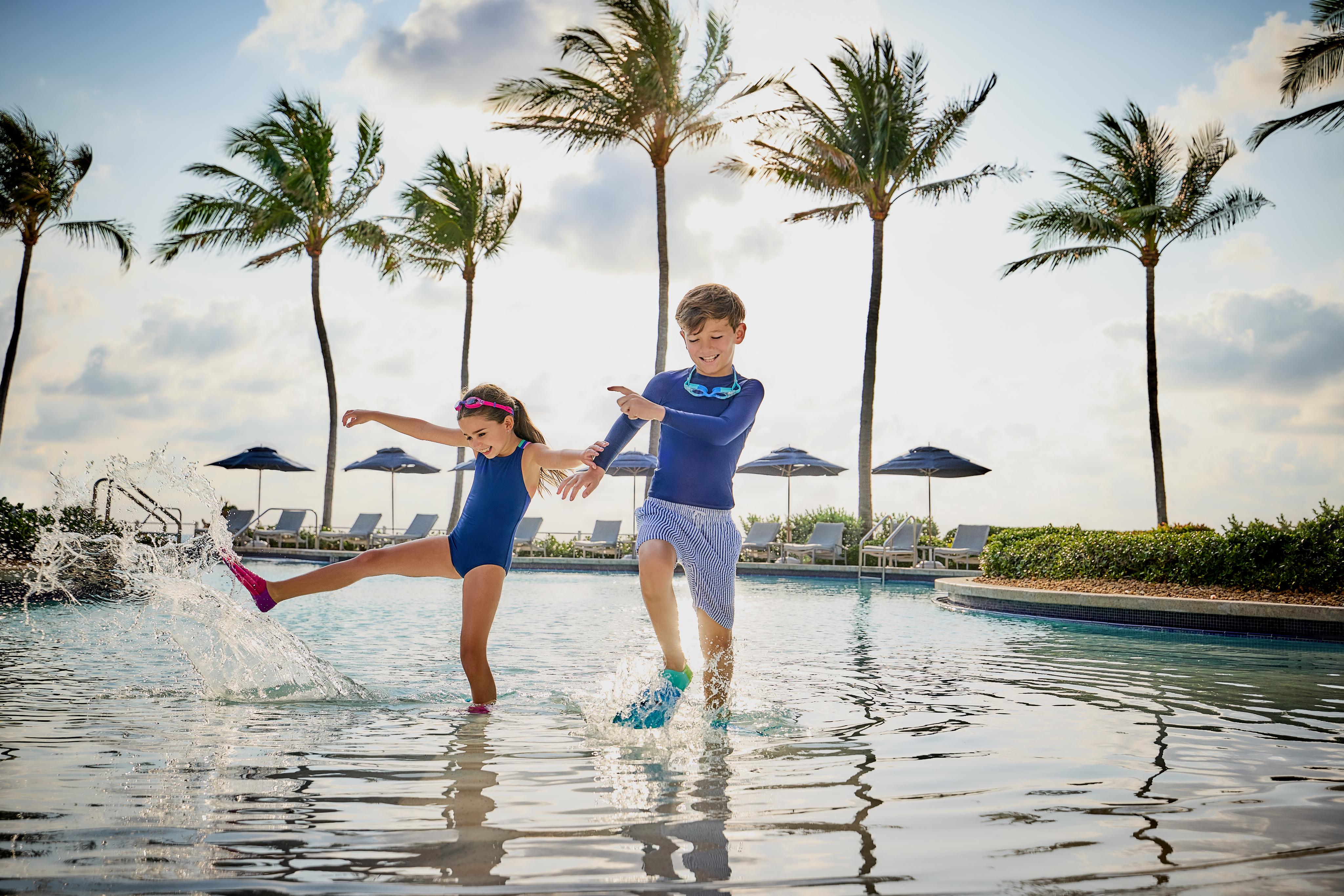 Two children in blue swimwear splashing and playing in a resort pool with palm trees in background.