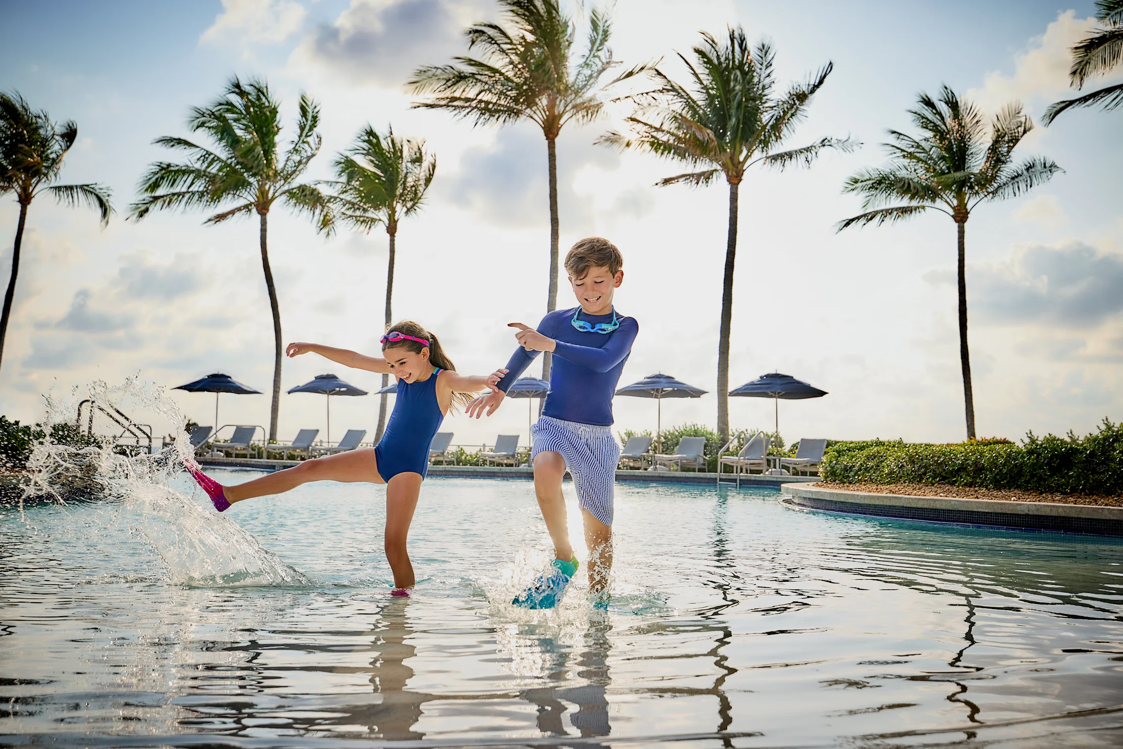 Two children in blue swimwear splashing and playing in a resort pool with palm trees in background.