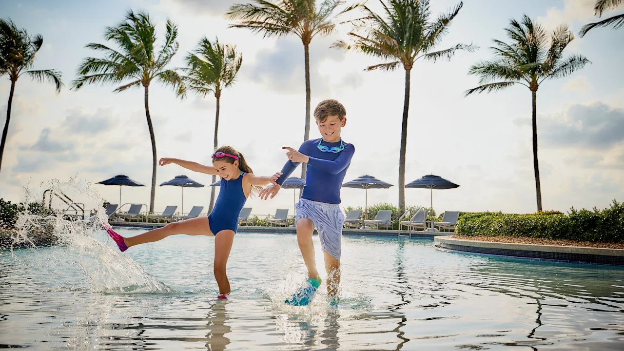Two children in blue swimwear splashing and playing in a resort pool with palm trees in background.