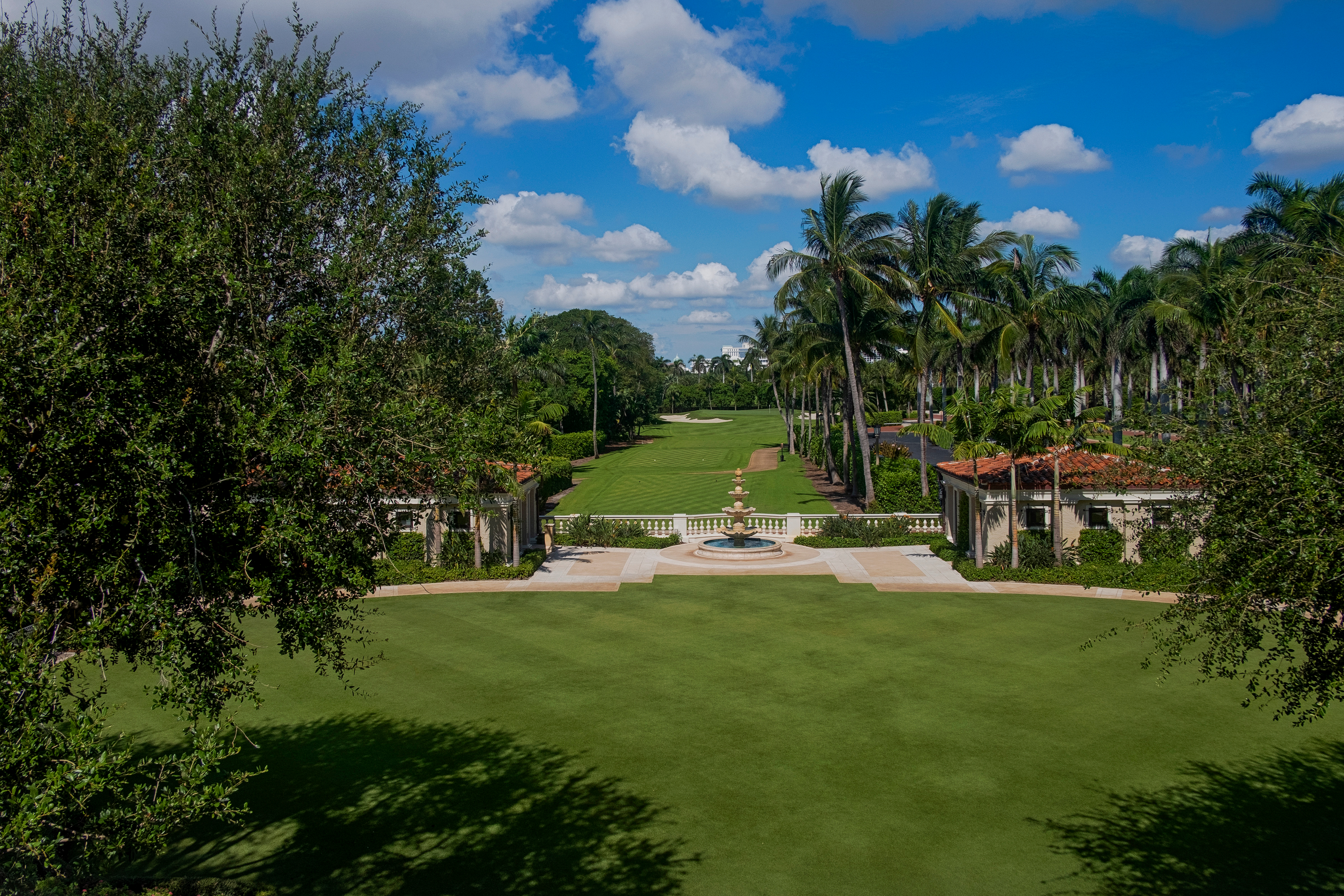 Manicured green lawn with palm trees, fountain, and Mediterranean-style buildings under blue sky.