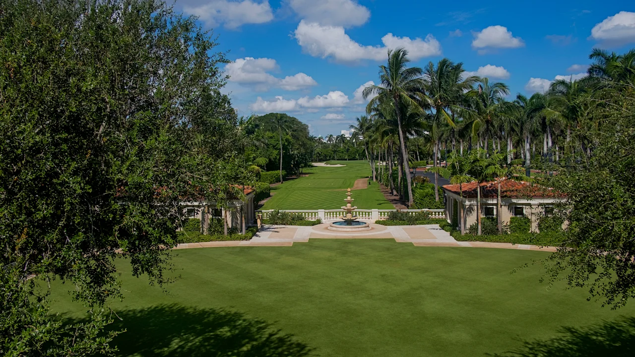 Manicured green lawn with palm trees, fountain, and Mediterranean-style buildings under blue sky.