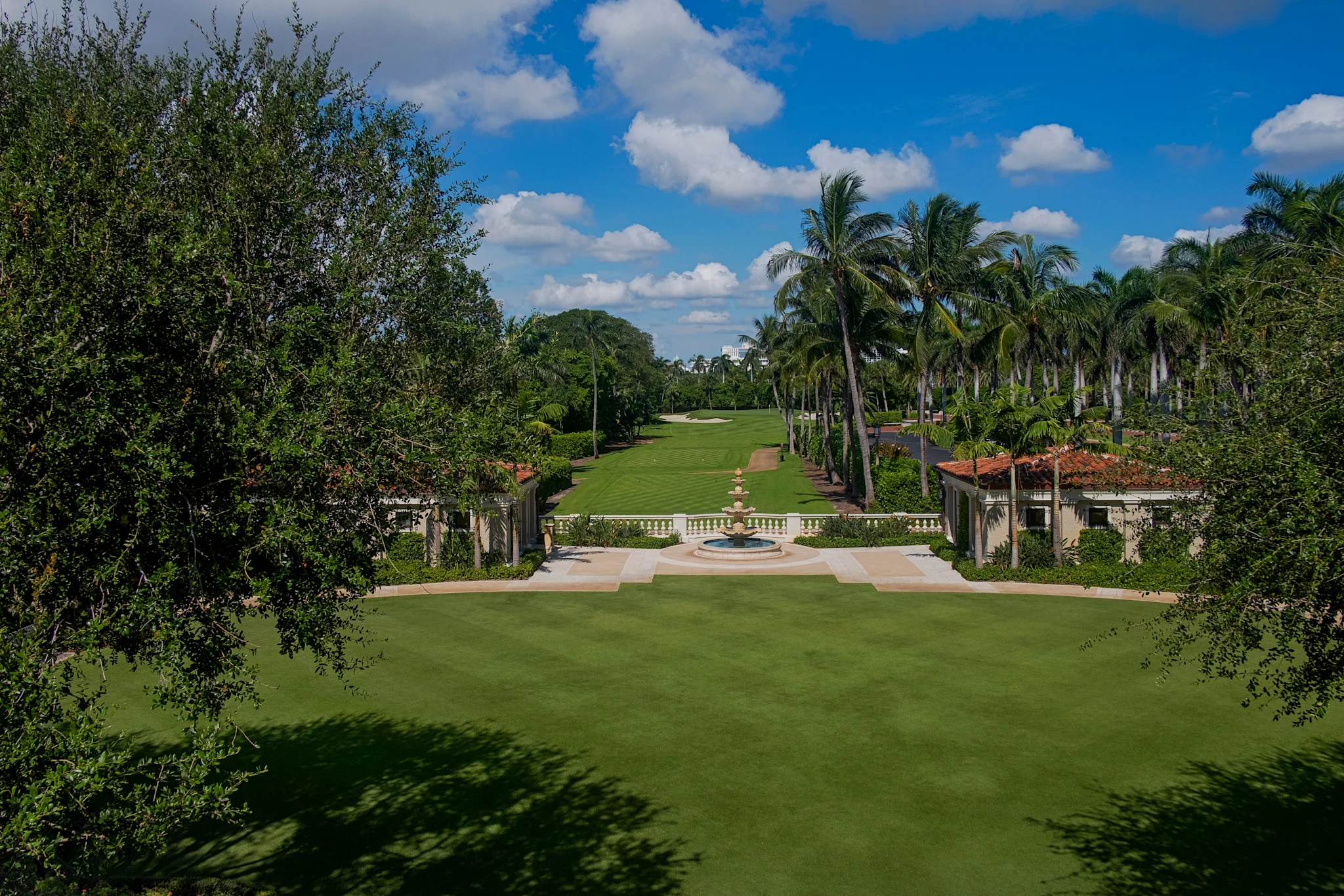 Manicured green lawn with palm trees, fountain, and Mediterranean-style buildings under blue sky.