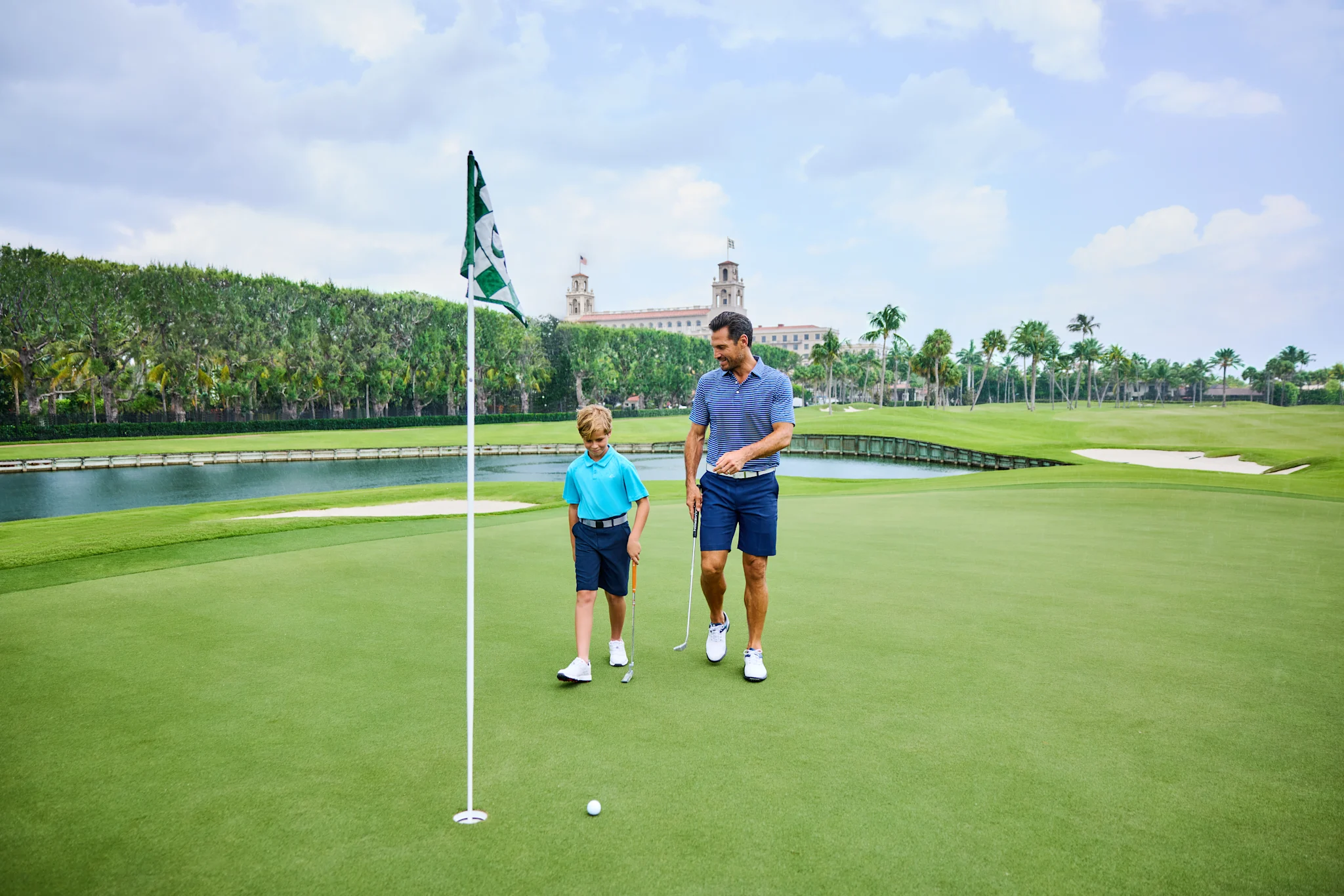 Father and son golfing on green course with water feature and historic building in background.