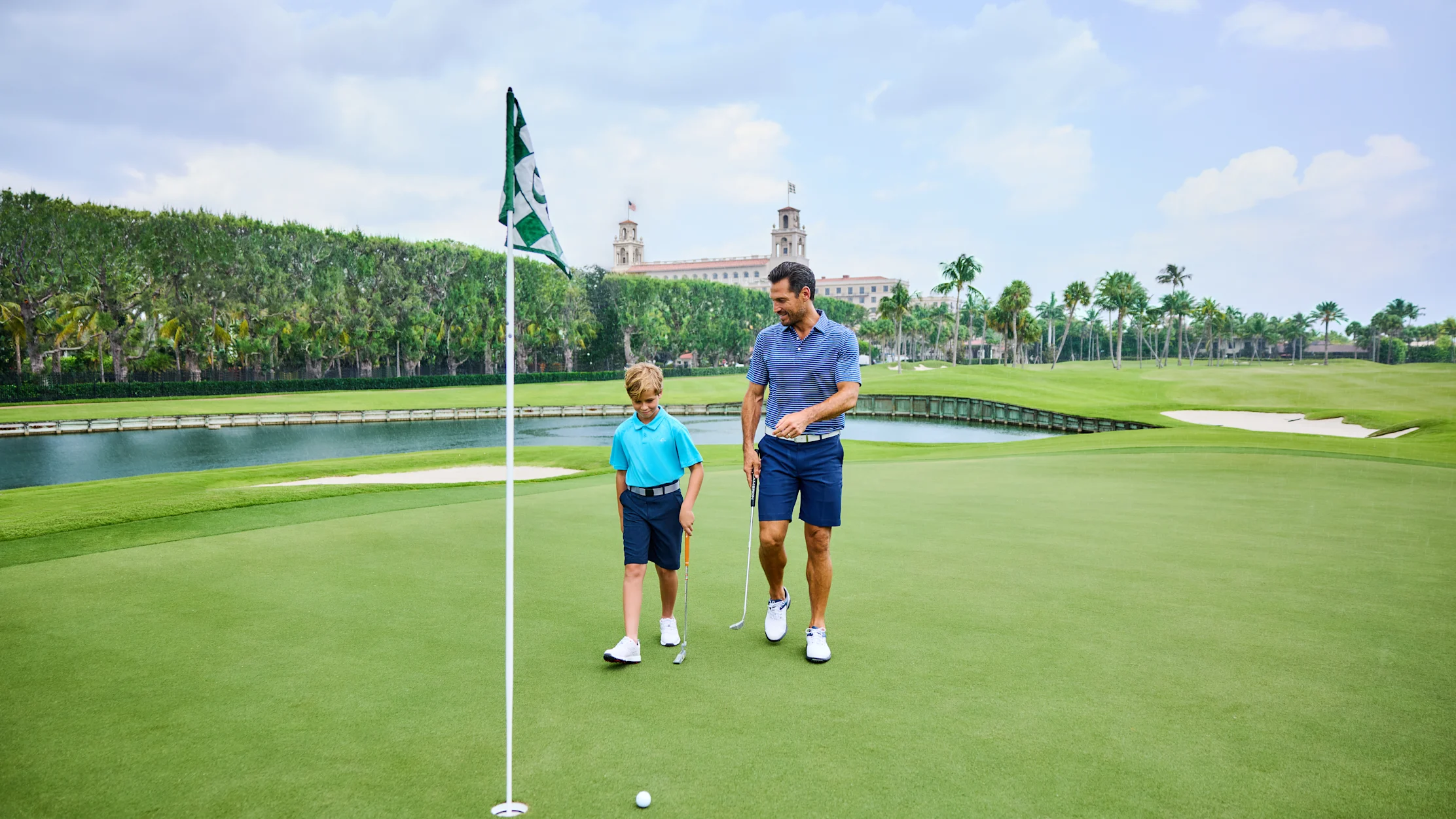 Father and son golfing on green course with water feature and historic building in background.