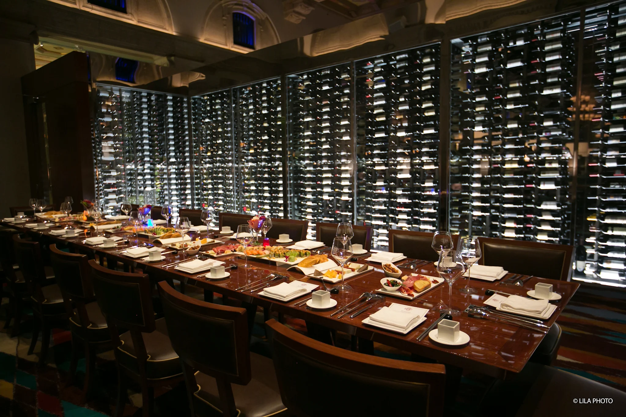 Elegant dining room with long wooden table set for dinner, backed by illuminated floor-to-ceiling wine racks.