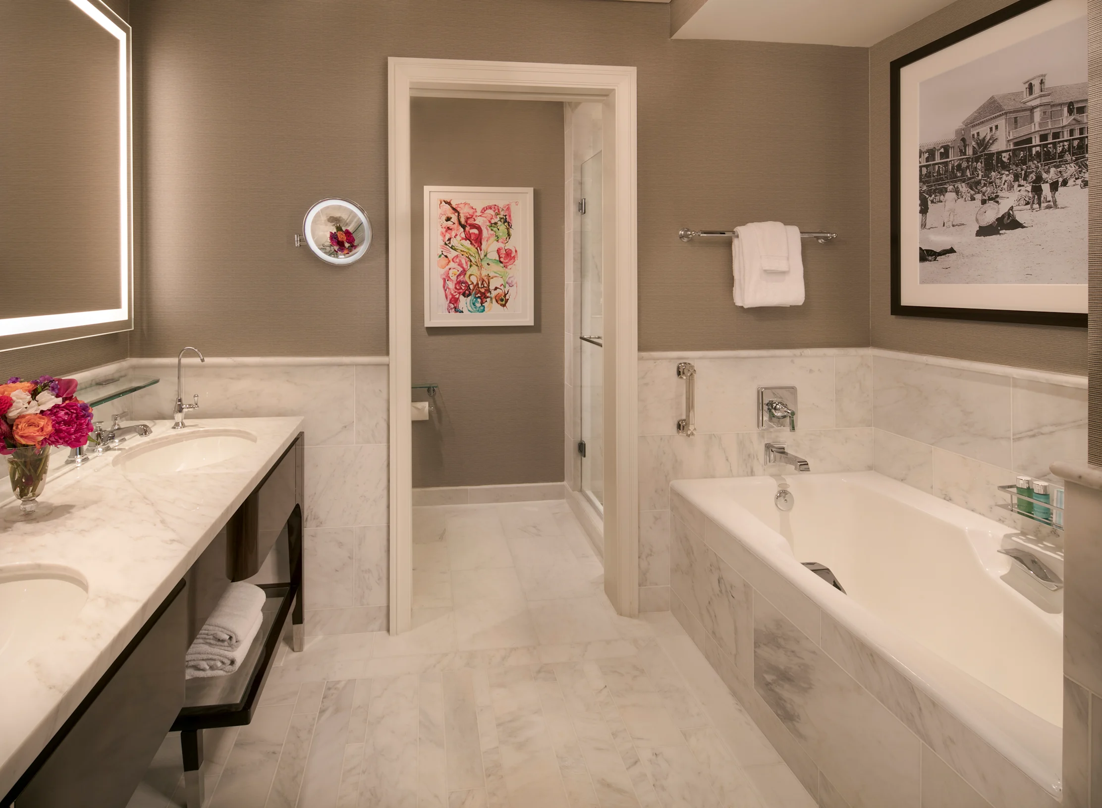 Elegant hotel bathroom with white marble, soaking tub, double vanity, and taupe walls with artwork.
