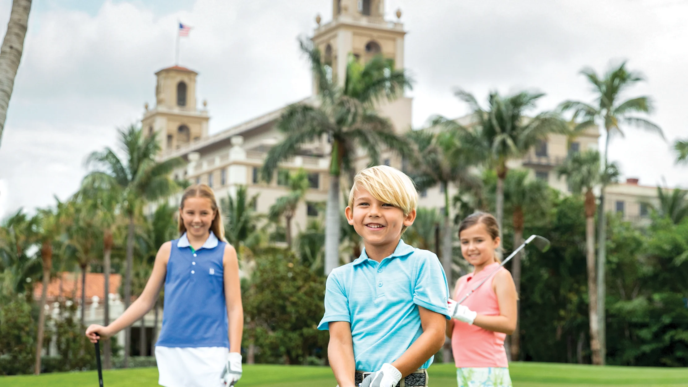 Three children with golf clubs on green course with palm trees and resort building in background