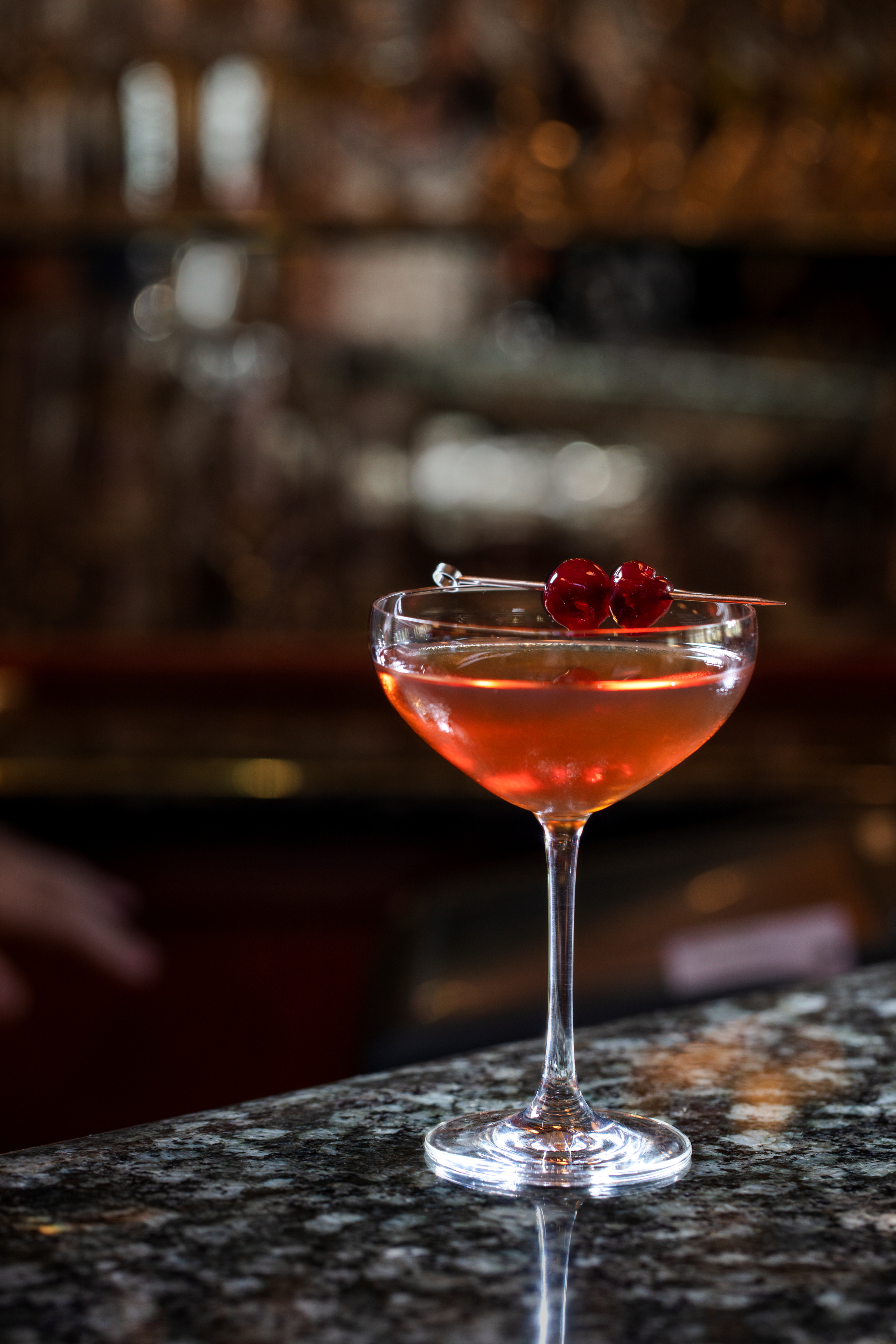 Orange cocktail in coupe glass garnished with red cherries on pick, bar background blurred