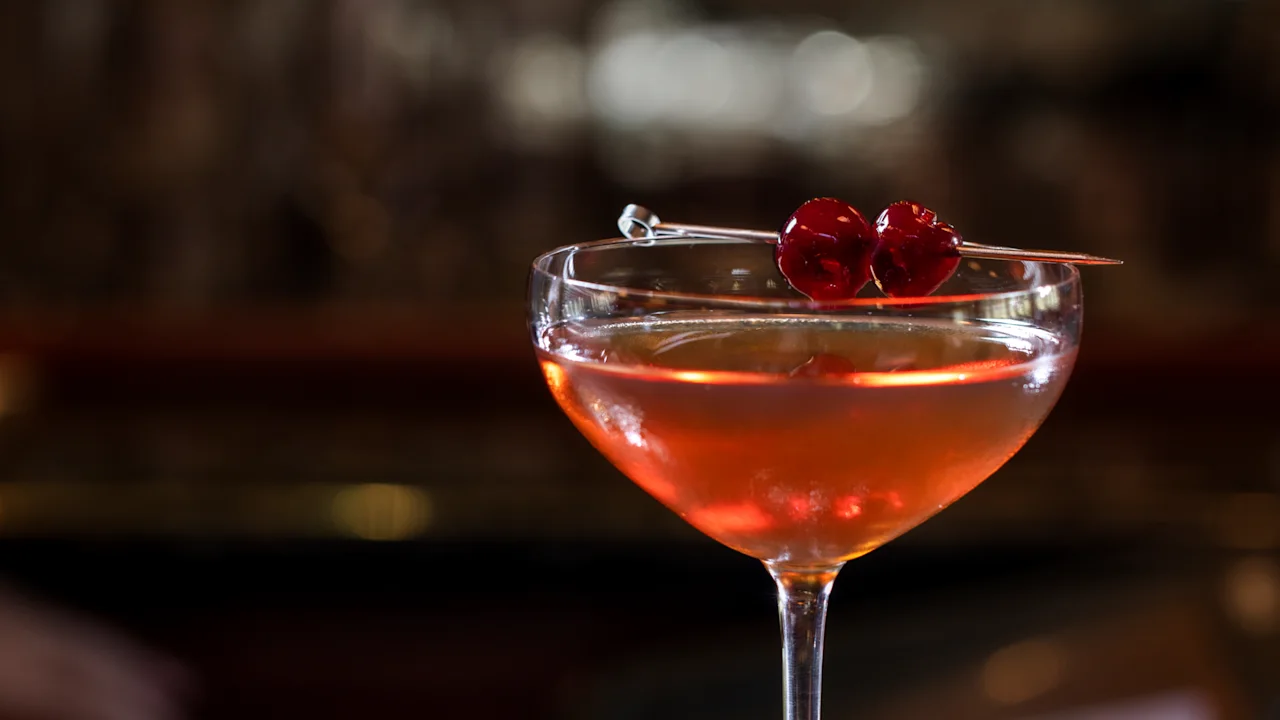 Orange cocktail in coupe glass garnished with red cherries on pick, bar background blurred