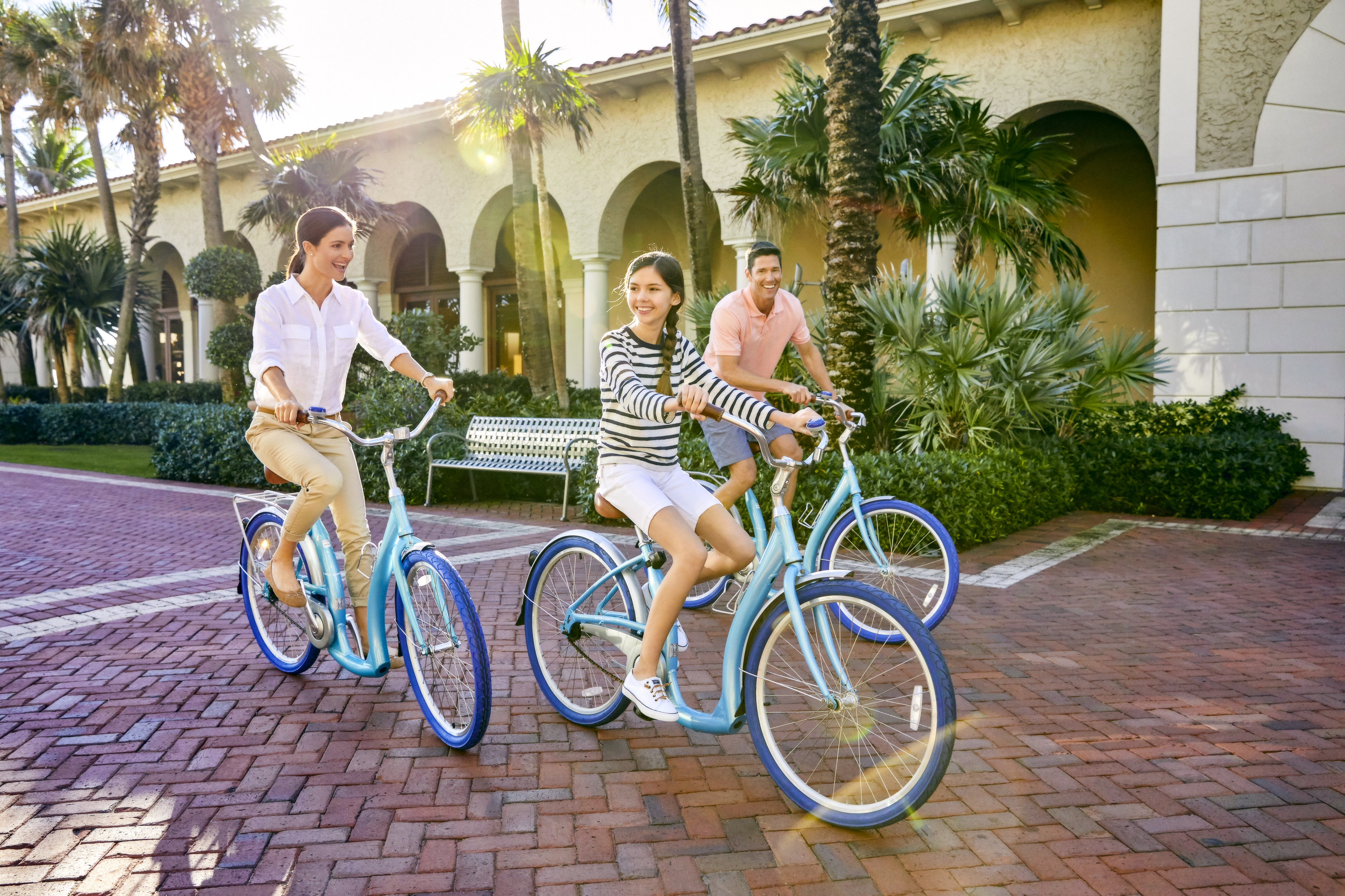Three people riding blue bicycles on brick pavement in front of a Mediterranean-style building.