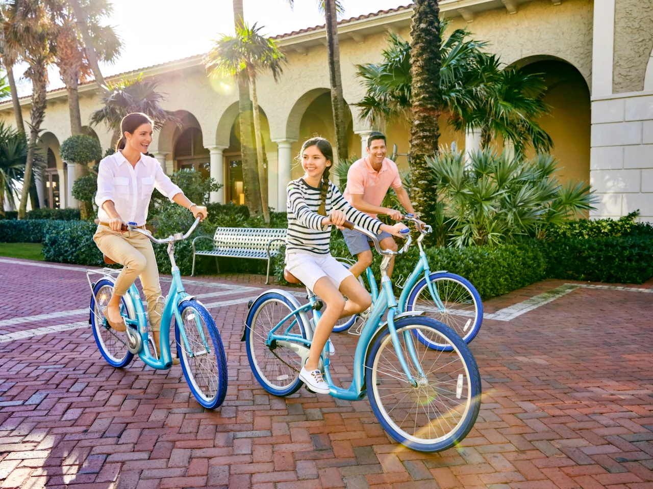 Three people riding blue bicycles on brick pavement in front of a Mediterranean-style building.