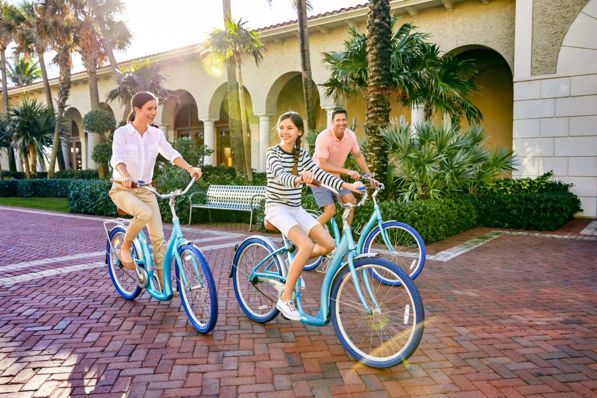 Three people riding blue bicycles on brick pavement in front of a Mediterranean-style building.