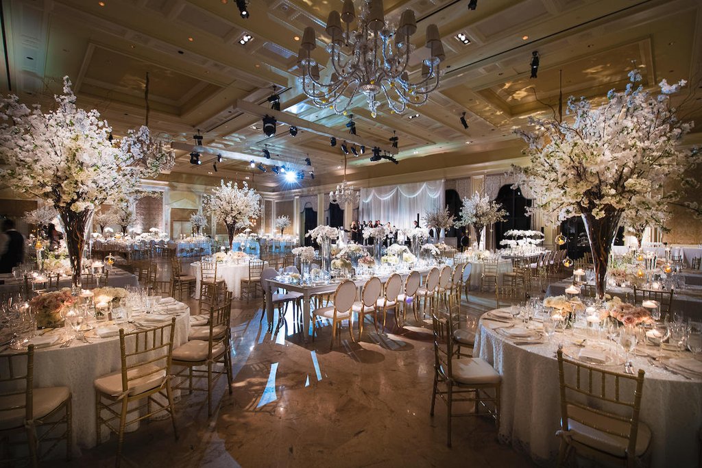 Elegant wedding reception ballroom with white floral centerpieces, crystal chandeliers, and gold chairs