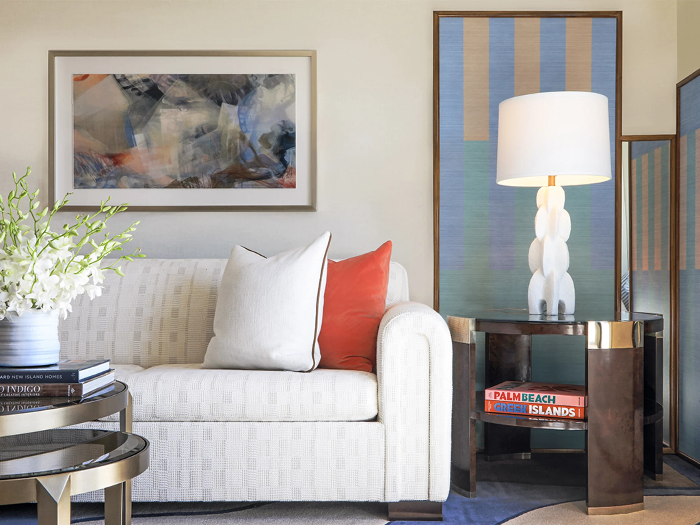 Modern living room with white sofa, coral pillow, abstract art, white table lamp, and striped artwork.
