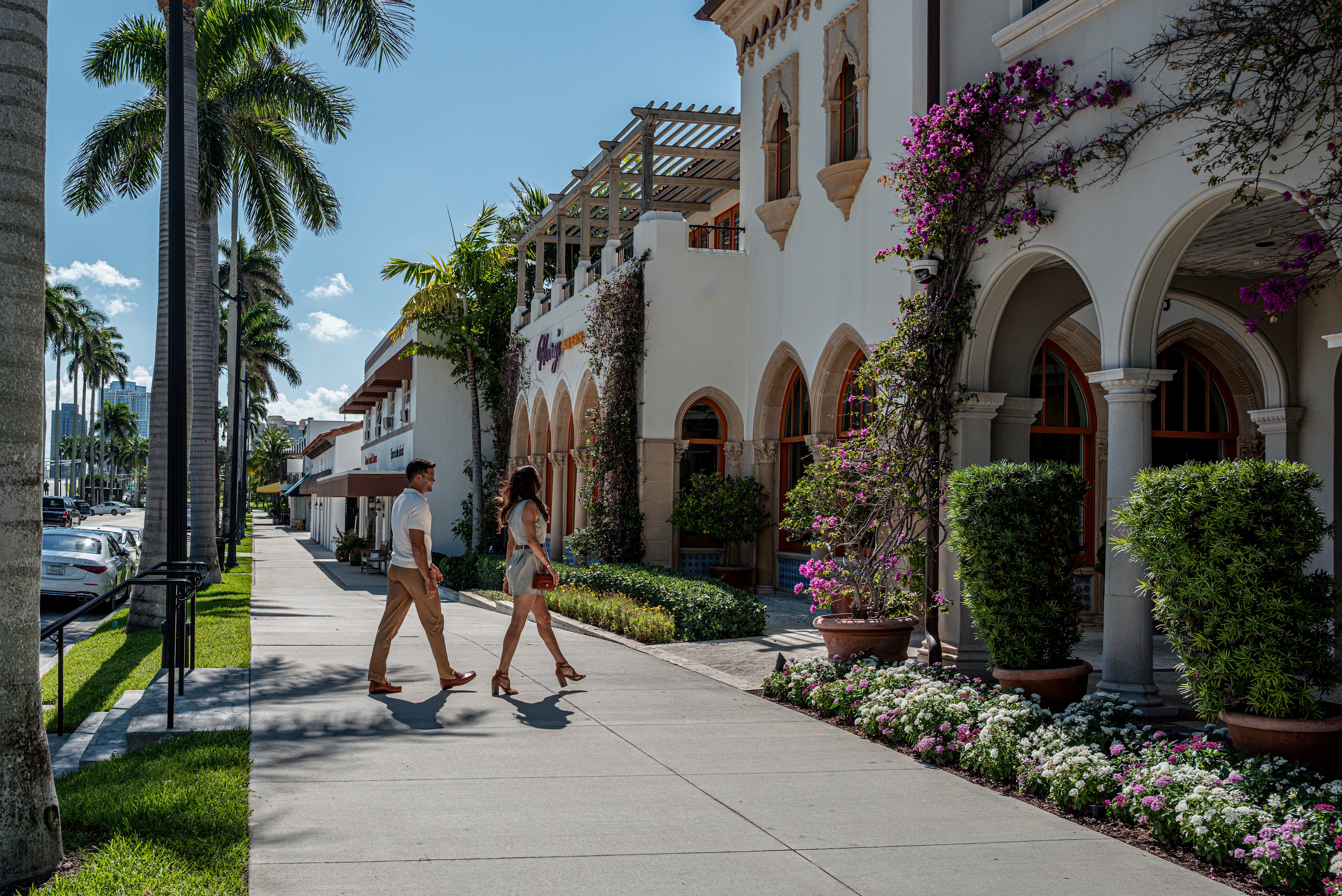 Couple walking past white Spanish-style building with arches, palm trees, and purple flowers.